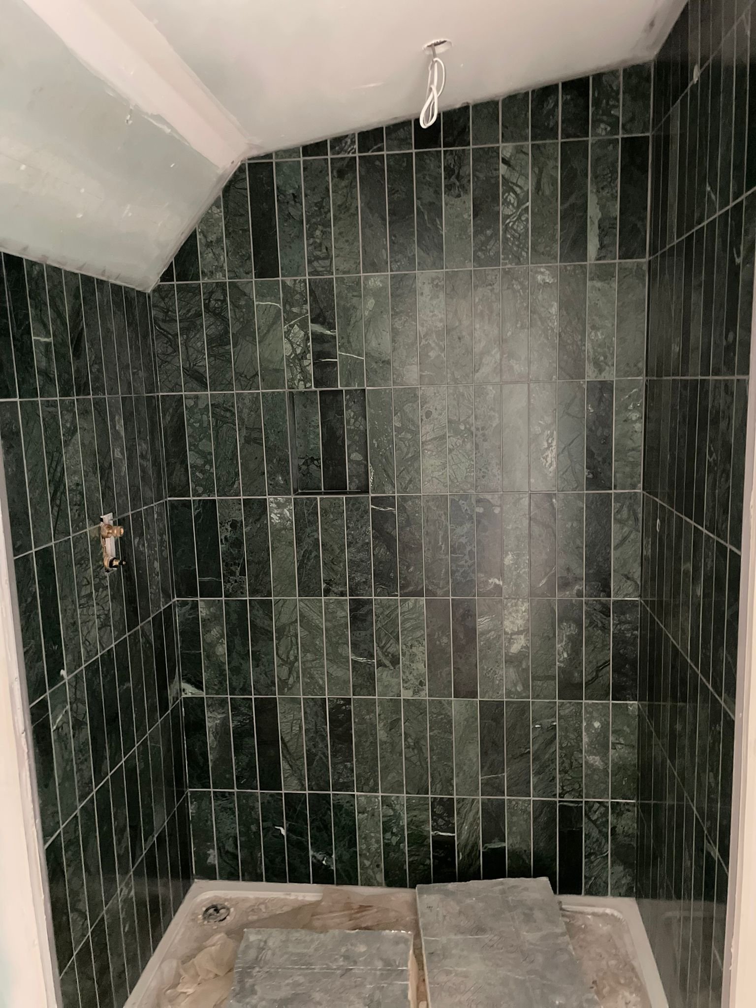 Empty shower with dark green tile walls, a ceiling with an exposed light fixture, and construction materials on the floor.