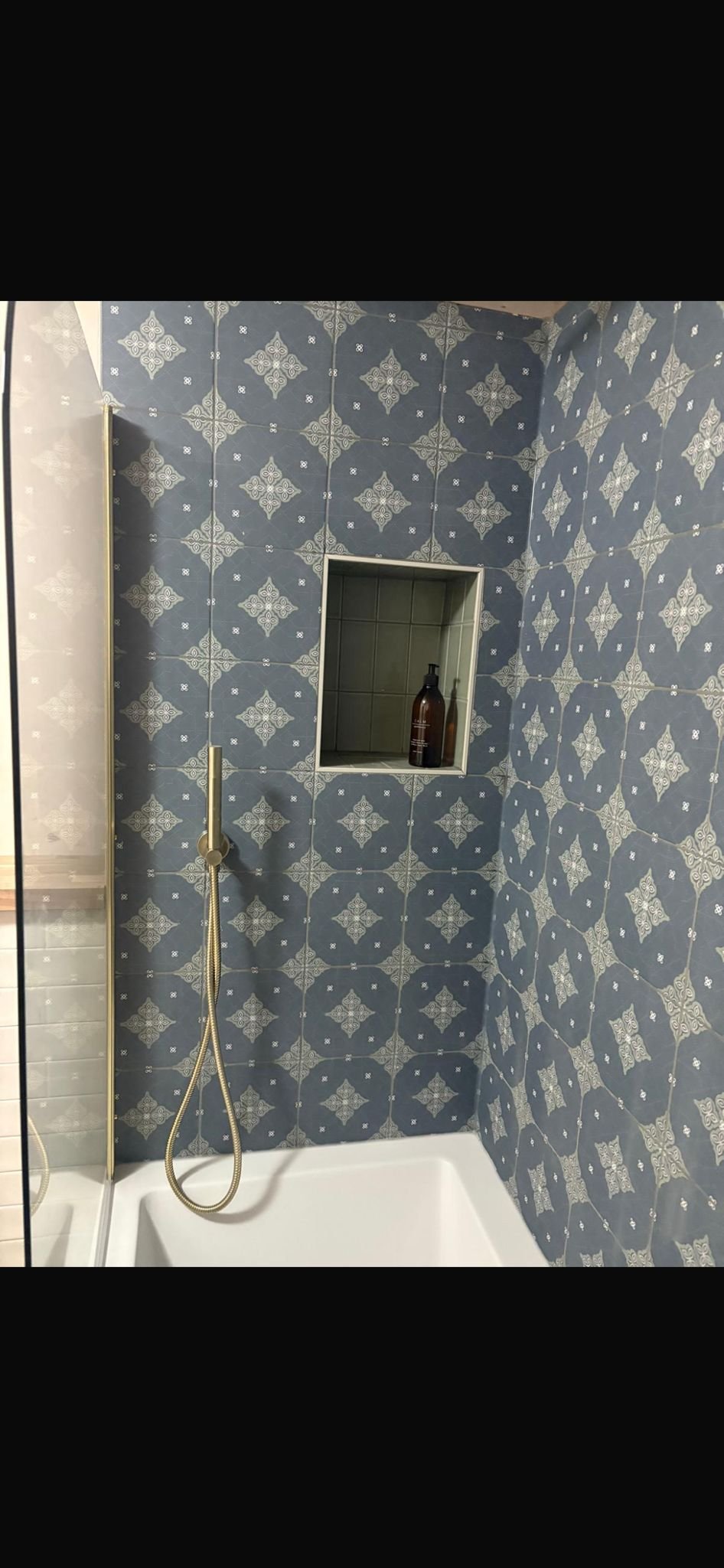 Bathroom shower with blue and white patterned tiles, a built-in wall niche with a brown bottle, a mounted shower head, and a partial view of a glass shower door.