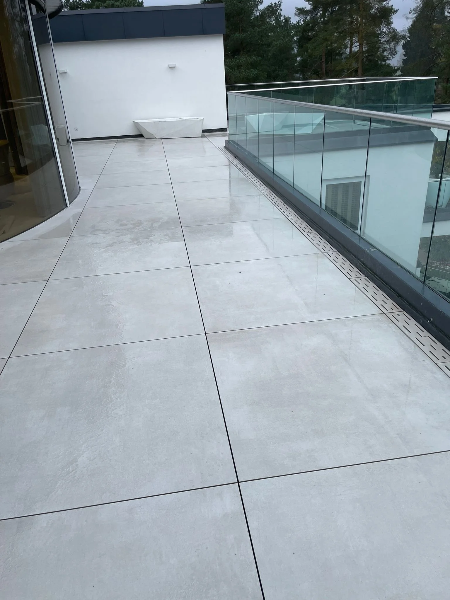 Modern balcony with large white tiled floor, glass railing, and a white wall with a decorative planter, overlooking trees.