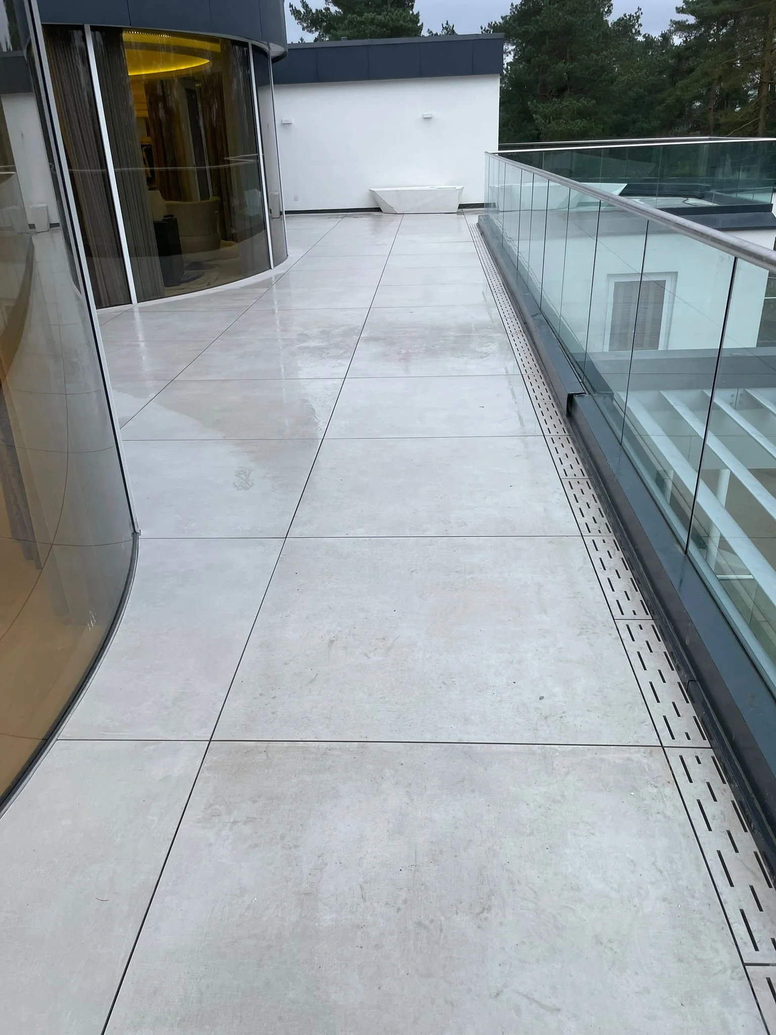 Modern balcony with light-colored tile flooring, glass railing, and a view of green trees in the background.