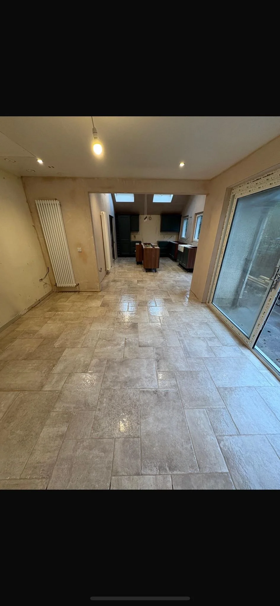 Empty room with tiled beige floor, white walls, radiator on left wall, large glass sliding door on right, and a kitchen area with cabinets and windows in the background.