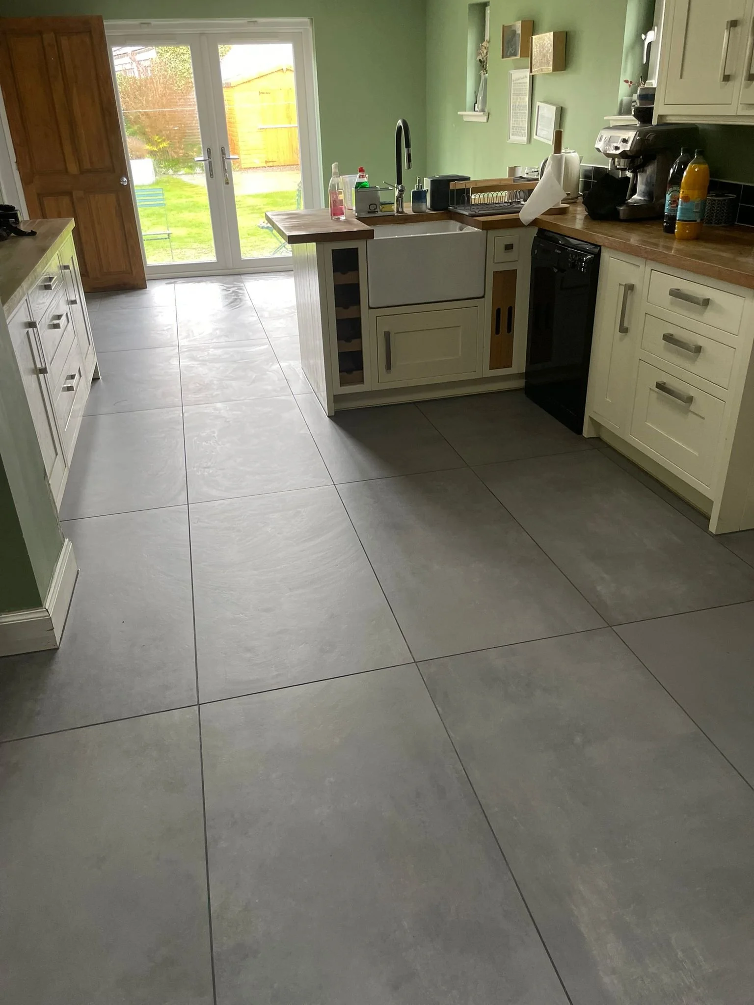 Bright kitchen with large gray tile floor, cream cabinets, a wooden countertop, and a green wall. There are sliding glass doors leading outside, with a small patio and yard visible.