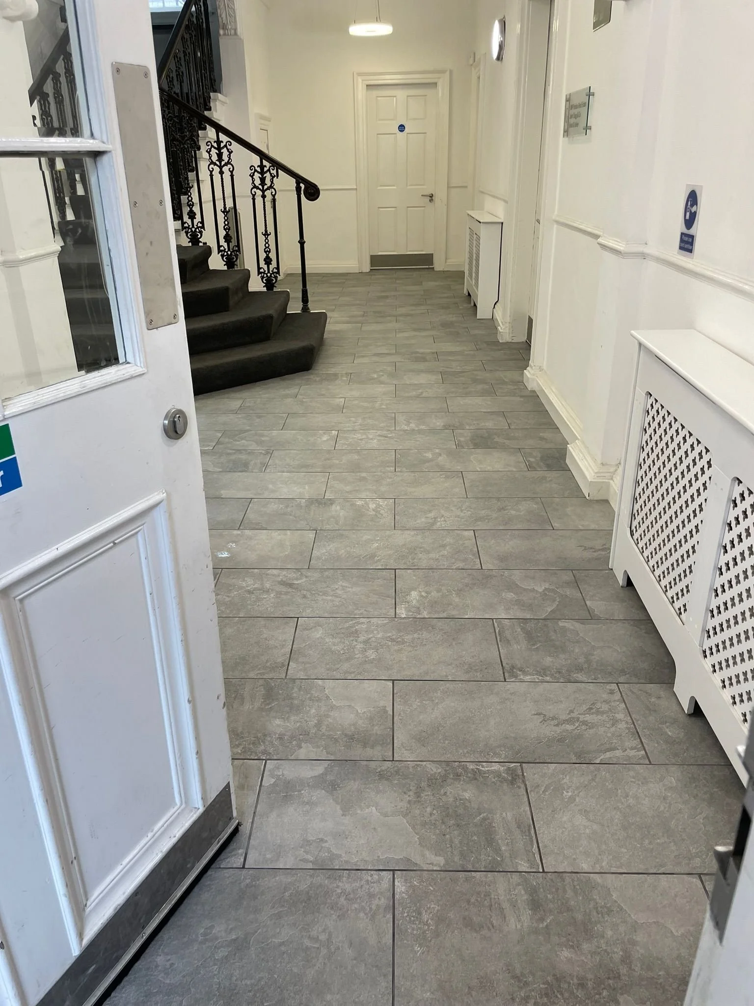 View of an indoor hallway with gray tiled flooring, white walls, and a staircase with black ornate railing on the left. A white radiator cover and a wall-mounted sign are on the right side, leading to a closed white door at the back.