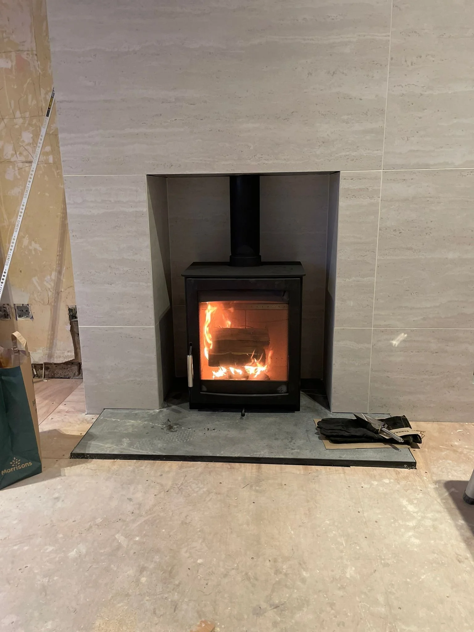 Black wood-burning stove with fire inside, installed in front of a beige tiled wall, with construction tools and materials nearby.