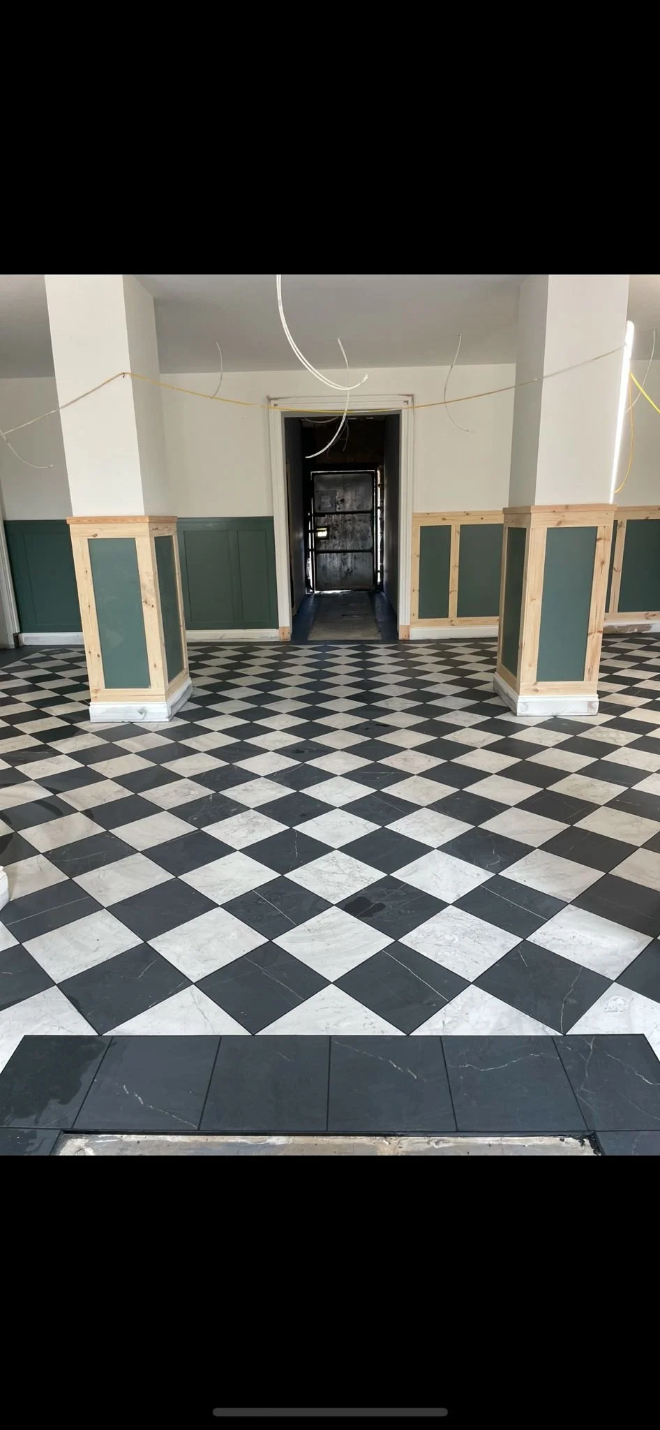 Interior space with black and white checkered tile floor, partially installed black tiles at the front, wooden wainscoting on the walls, and hanging wires from the ceiling, possibly under construction or renovation.
