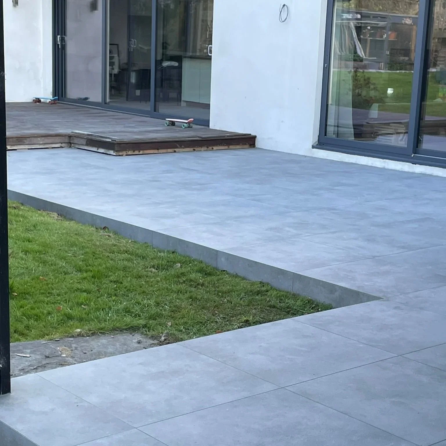 A modern house patio with grey tiled floor, a small patch of green grass, and a sliding glass door leading inside. There are skateboards on the wooden deck.