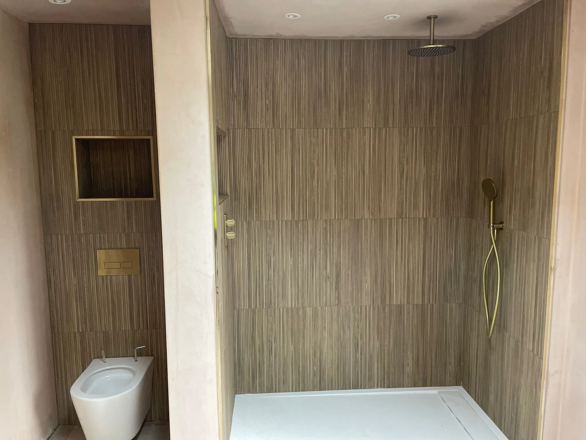View of a modern bathroom with a shower area on the right, featuring a ceiling-mounted showerhead and a handheld shower on the wall, and a white bidet on the left side below a wooden wall panel with a rectangular niche.