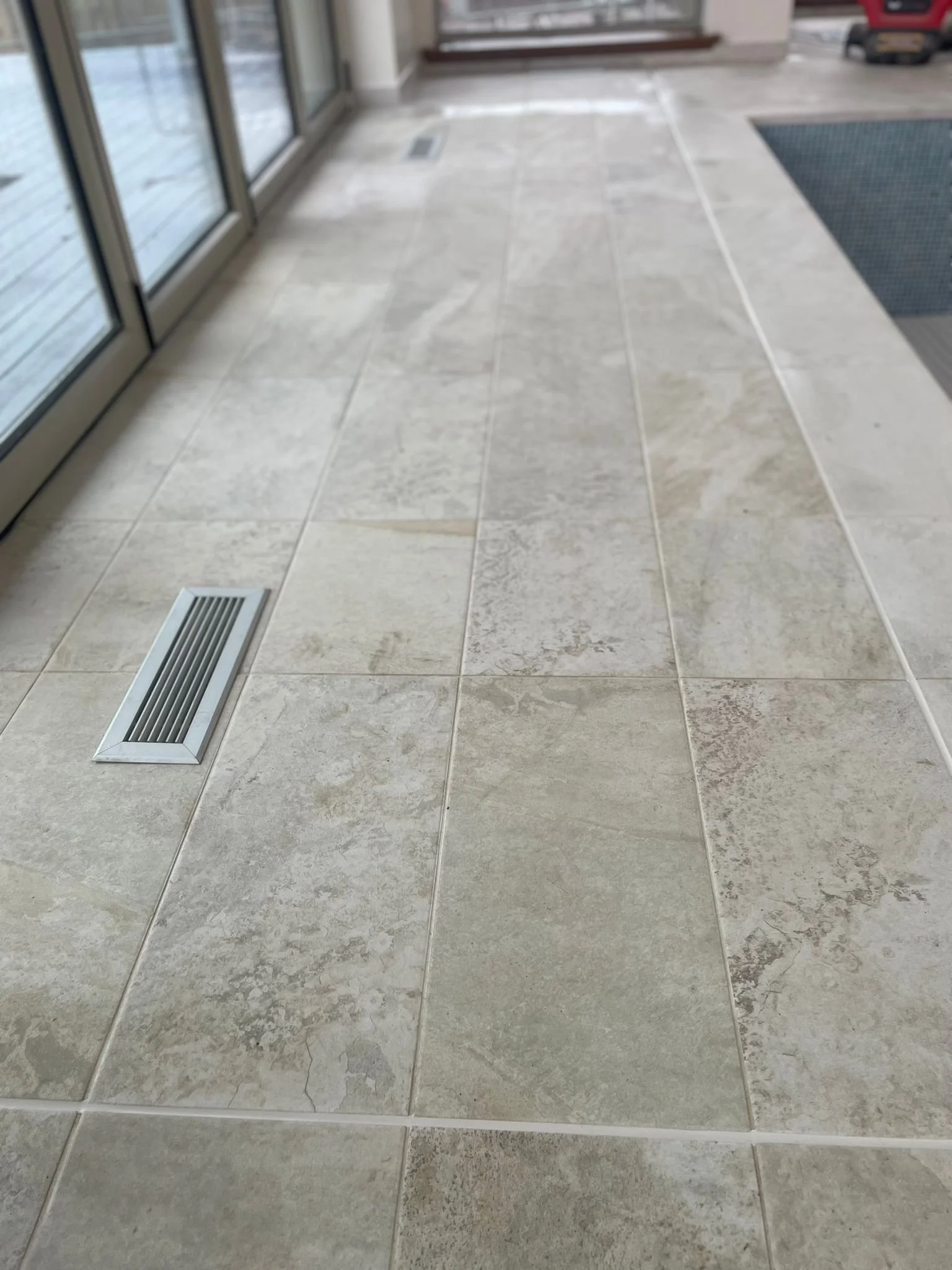 Indoor tiled floor near glass doors, with a metal vent and a doormat in the background.
