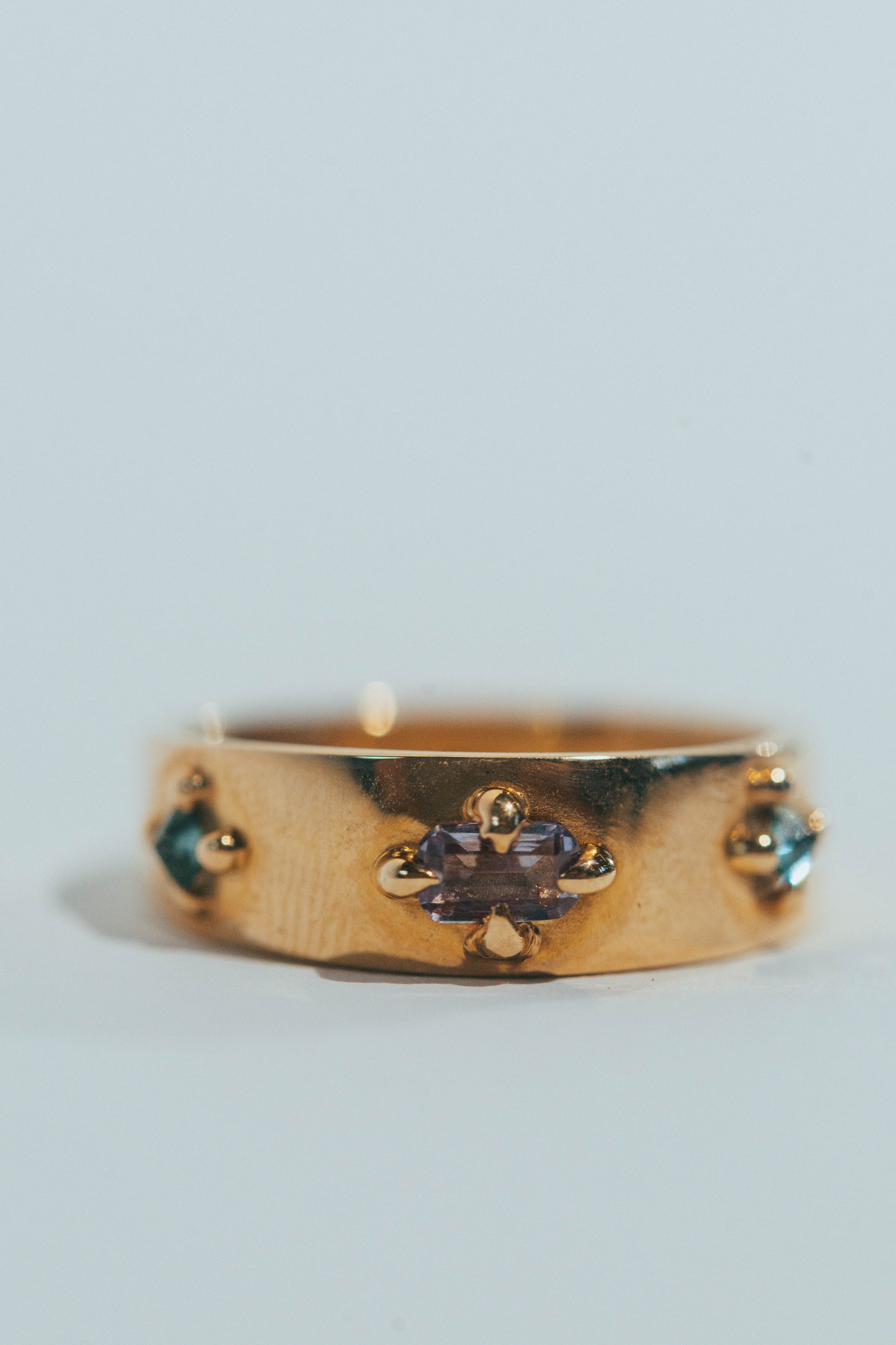 Close-up of a gold ring with multiple gemstones, including a central purple stone and smaller blue stones on each side.