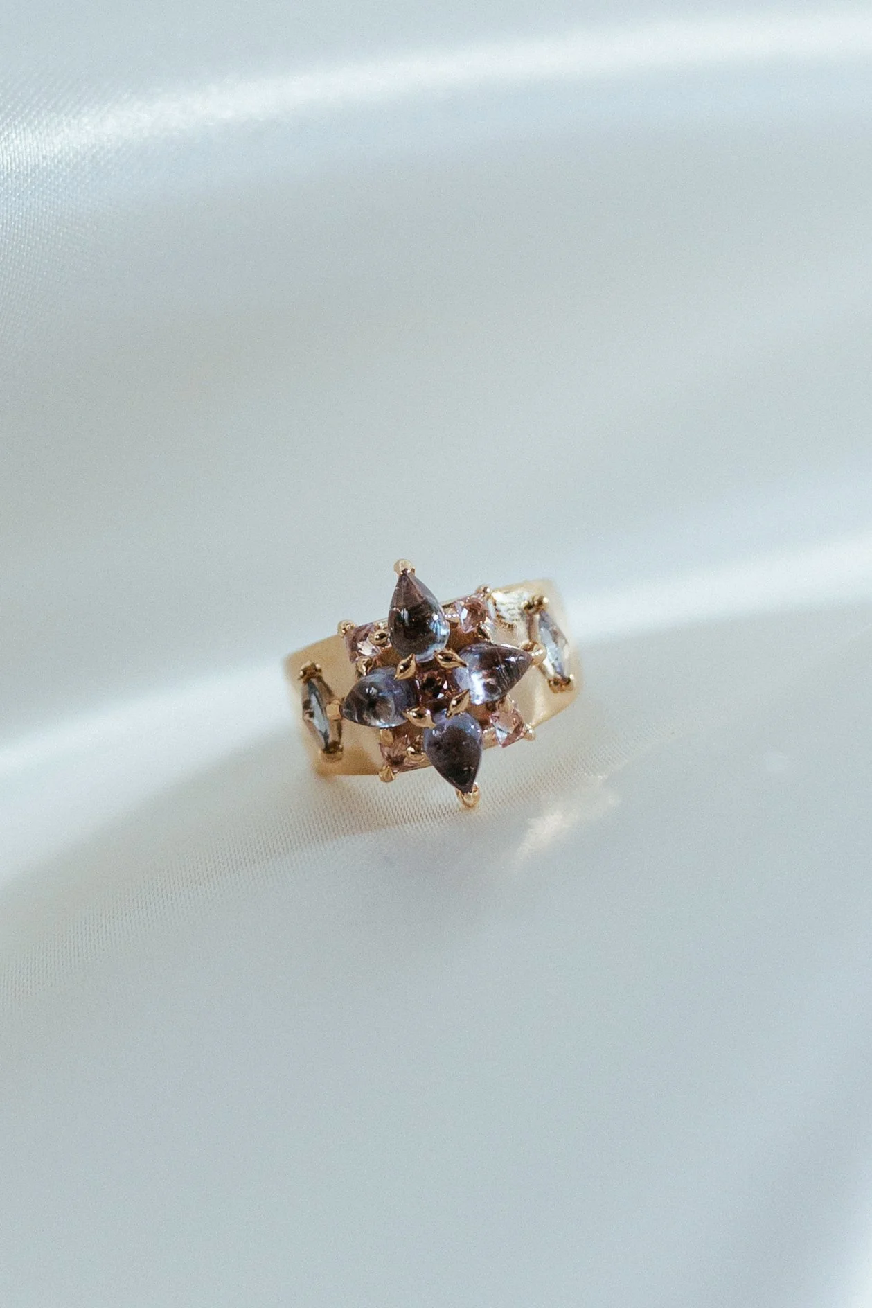 Close-up of a gold ring with dark purple and clear gemstones on a satin fabric surface.