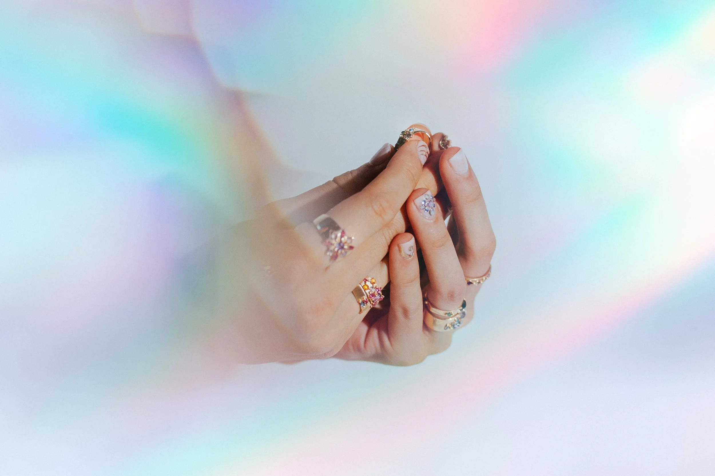Close-up of multiple hands with various rings on fingers, holding a ring between thumb and forefinger, against a pastel, iridescent background.