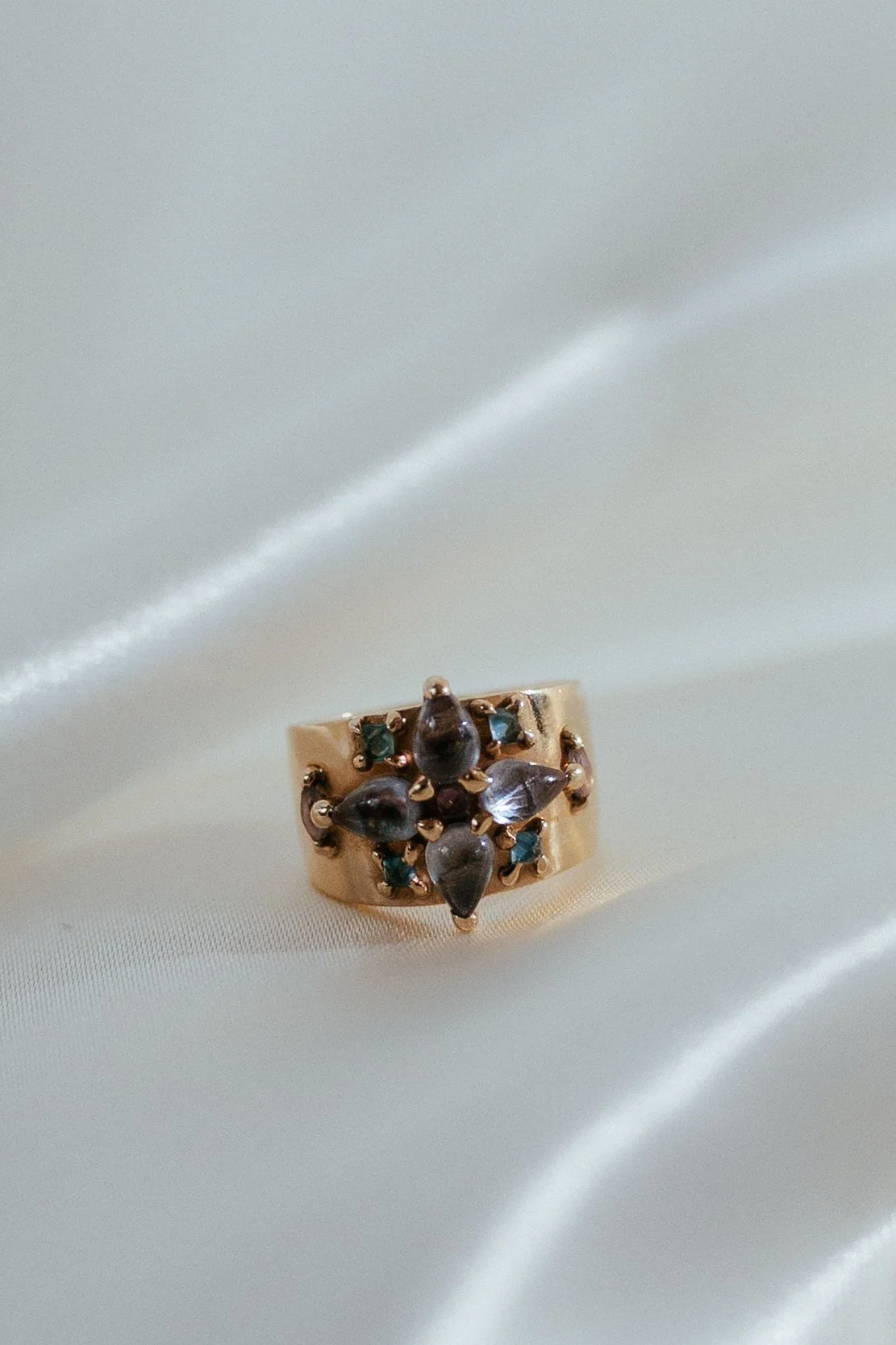 A gold ring with black and blue gemstones resting on smooth white satin fabric.