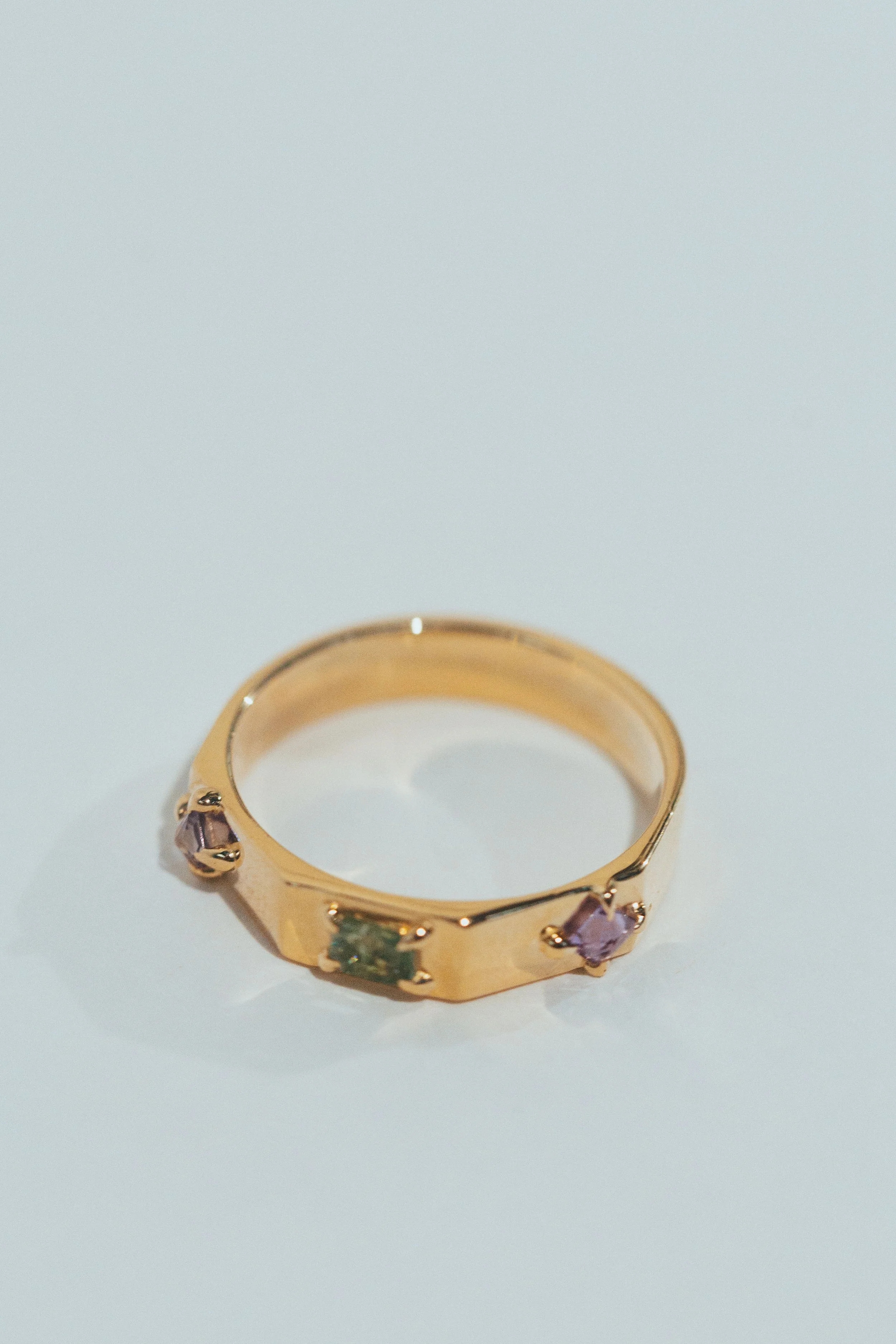 Gold ring with three gemstones, including purple and green stones, on a white surface.