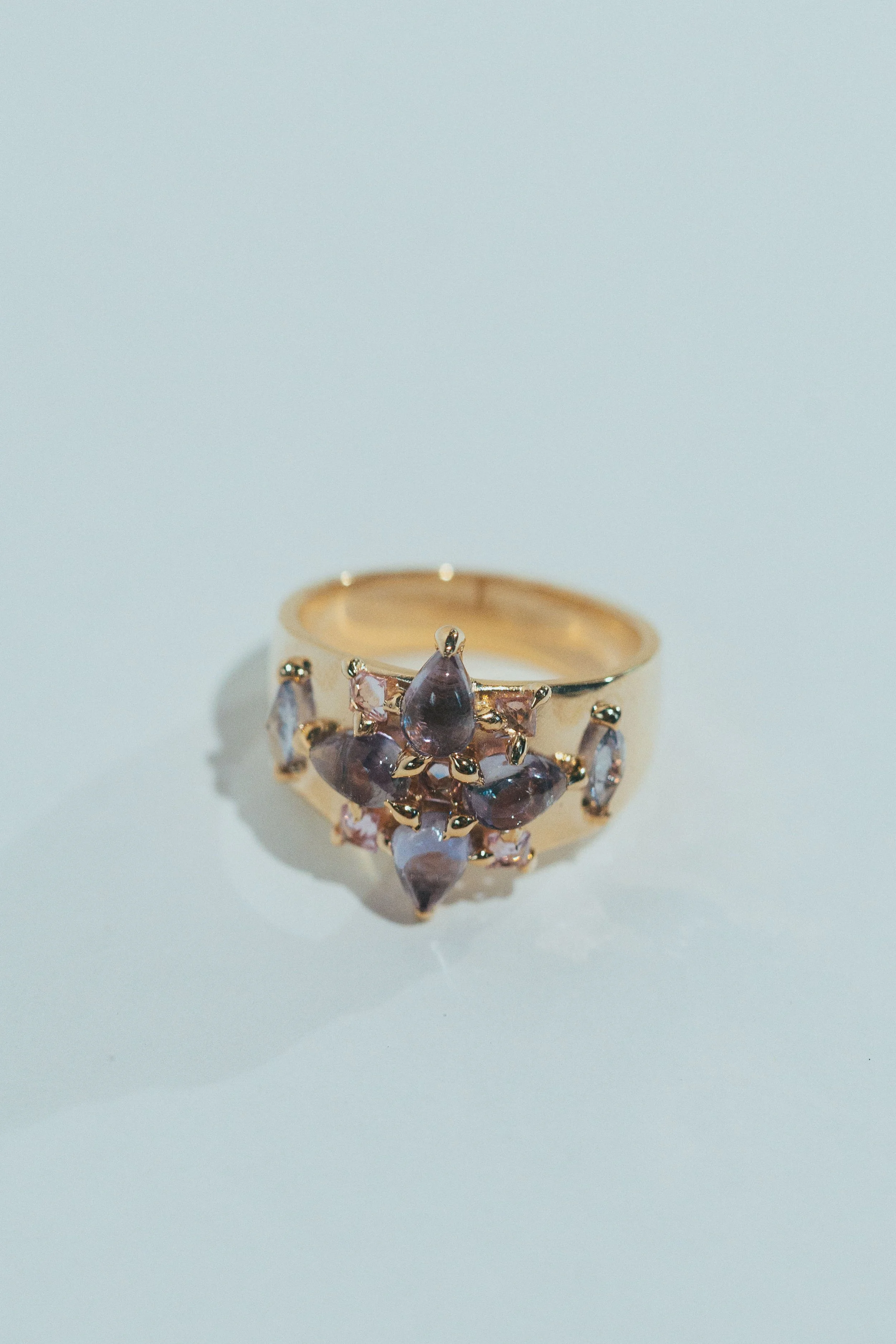 Gold ring with purple and pink gemstones arranged in a floral pattern on a white surface.