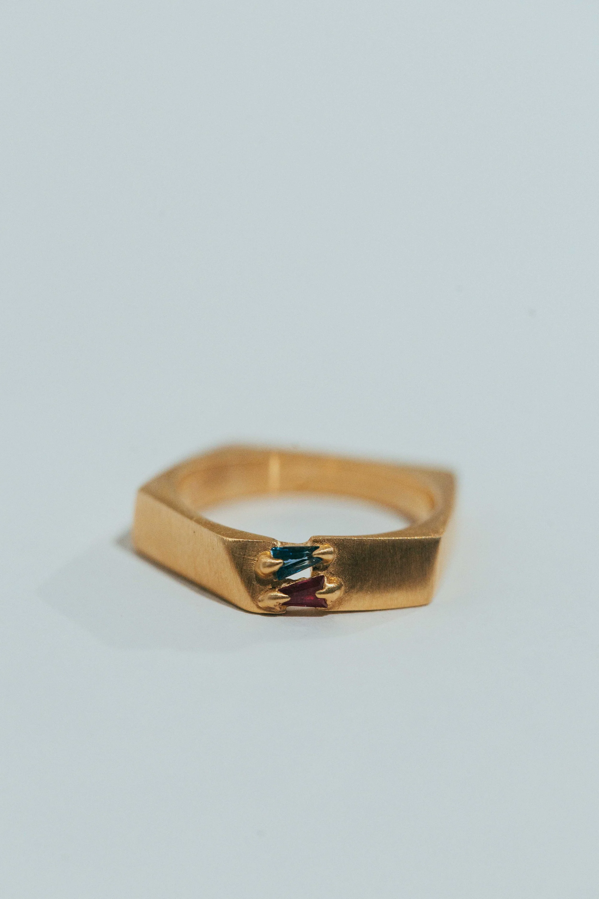 A gold ring with a geometric design featuring two small colored gemstones, one blue and one red, set on top.