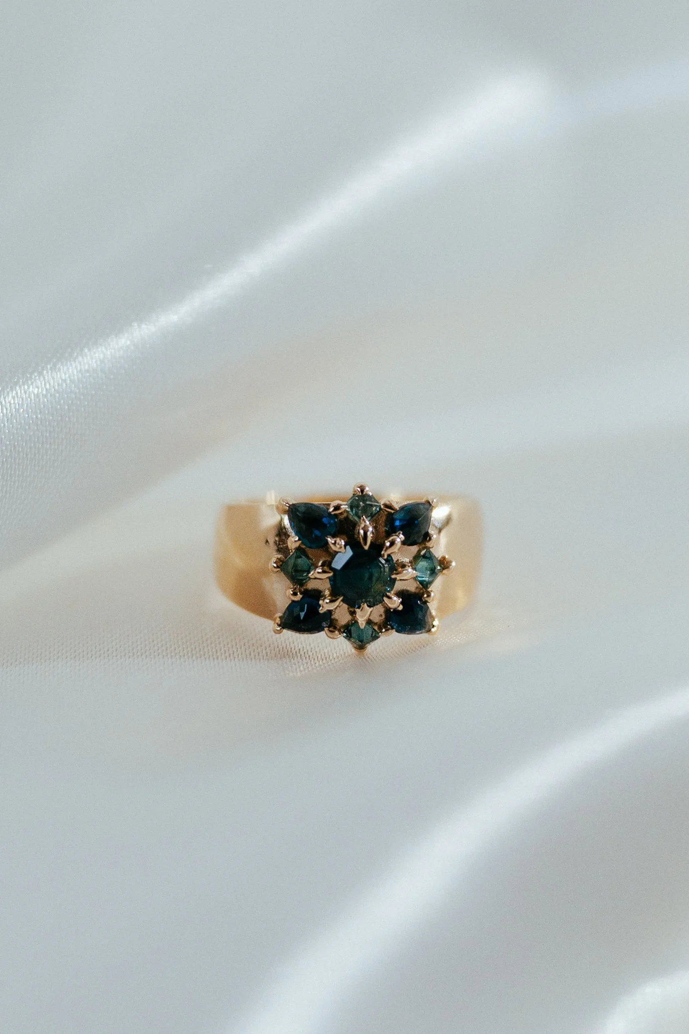 A gold ring with a cluster of dark blue and green gemstones on a white satin fabric.