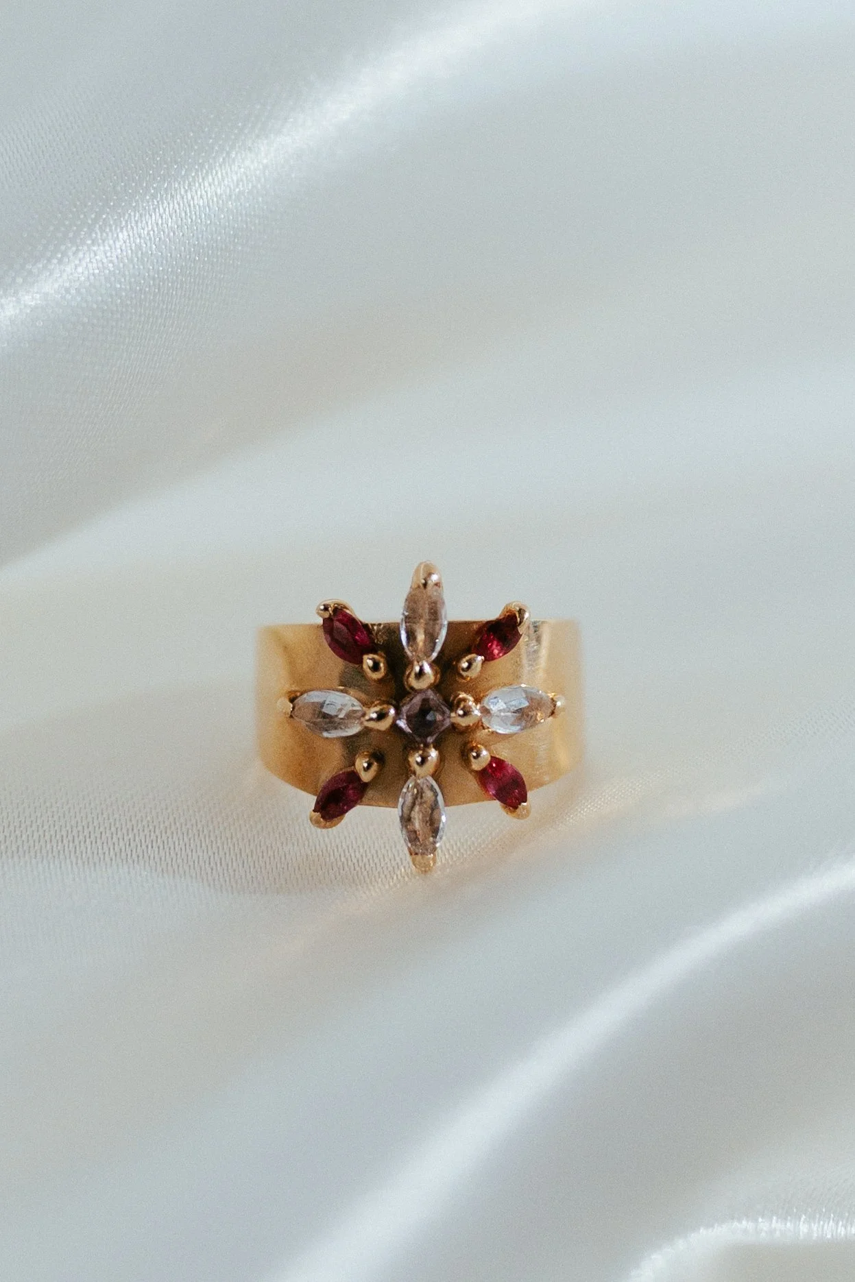 Gold ring with a floral design of marquise-cut red and clear gemstones, set on white satin fabric.