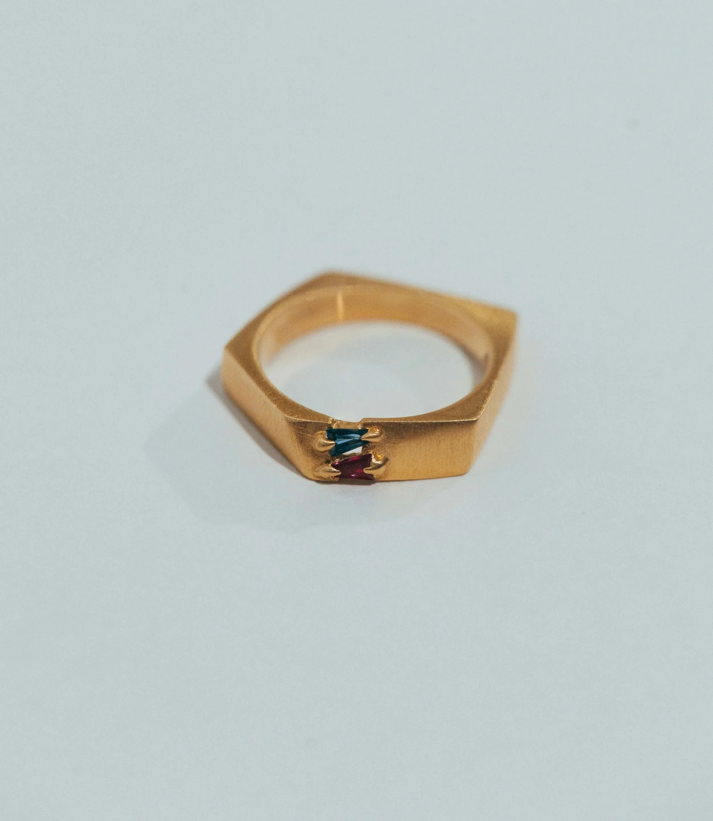 Gold ring with two small gemstones, one blue and one red, embedded on the side, placed on a white surface.