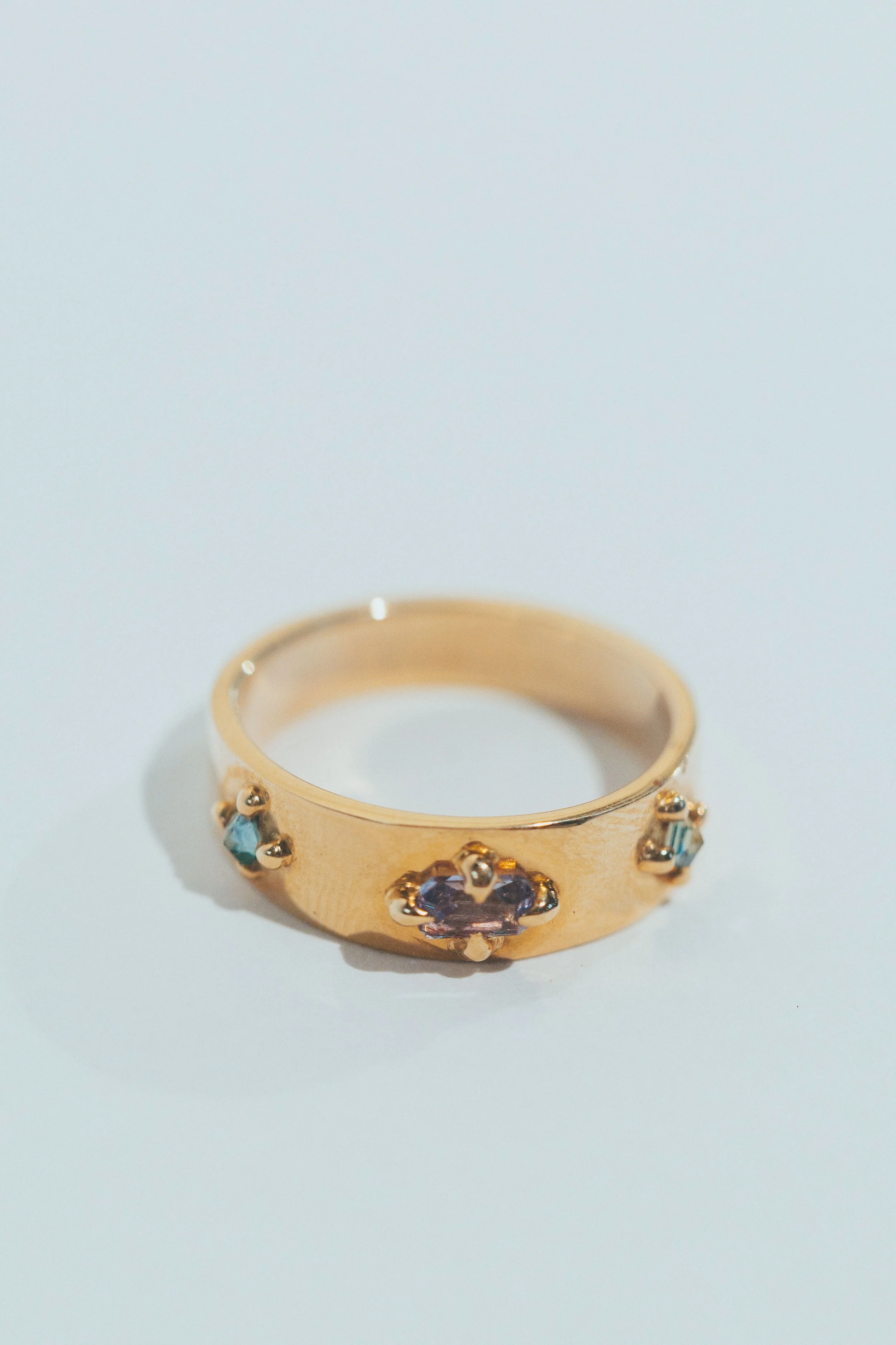 Gold ring with three small gemstones, one purple and two blue, set apart on a plain white background.