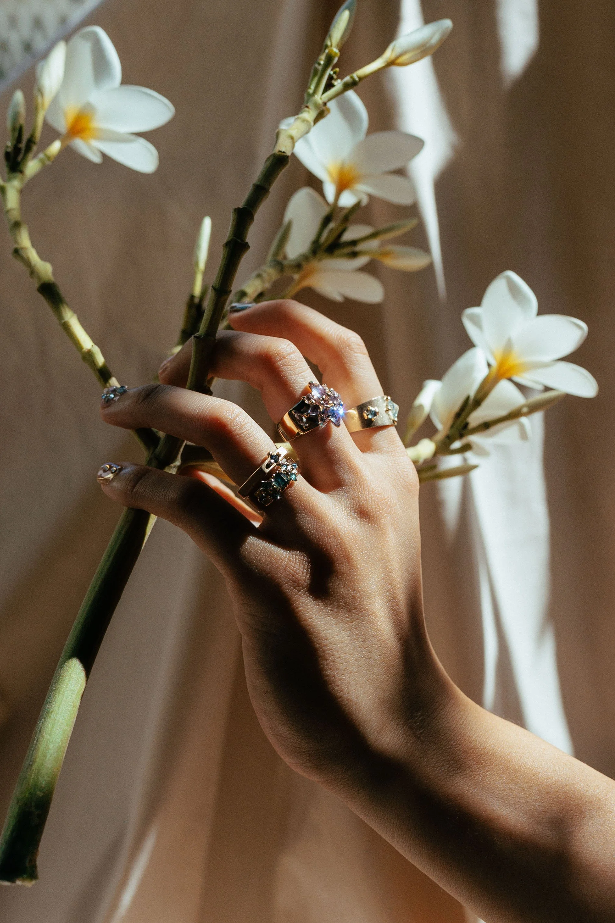 A person's hand holding a branch of white plumeria flowers, with multiple rings adorned with gemstones on their fingers, in natural lighting. 14kt yellow gold and sapphires. Handmade in Hawaii. One-of-a-kind Jewelry. Bridal, engagement rings.