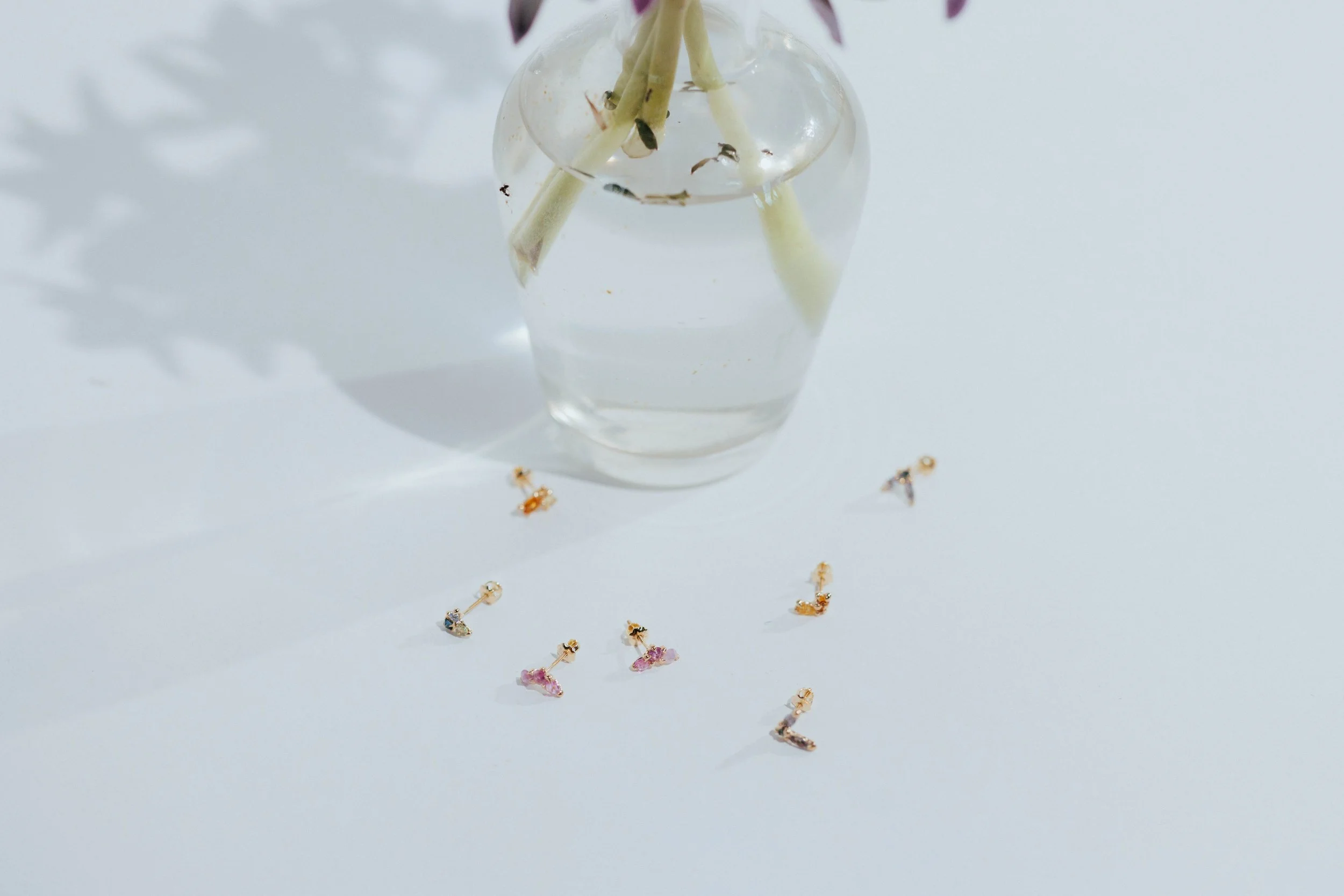Clear glass vase with water and white flowers, placed on a white surface, with scattered small colorful earrings around it.