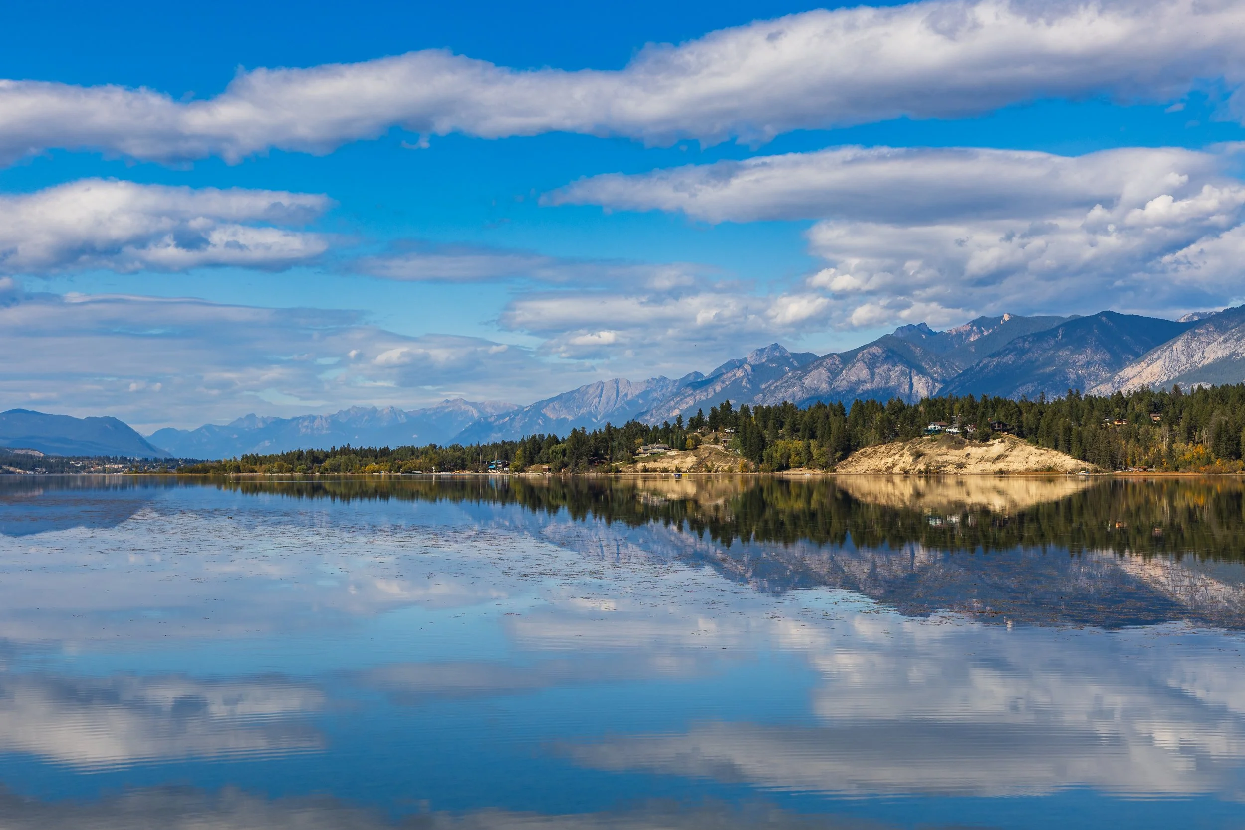 Scenic view of a lake with calm, reflective water, surrounded by forested hills and mountains under a partly cloudy sky.
