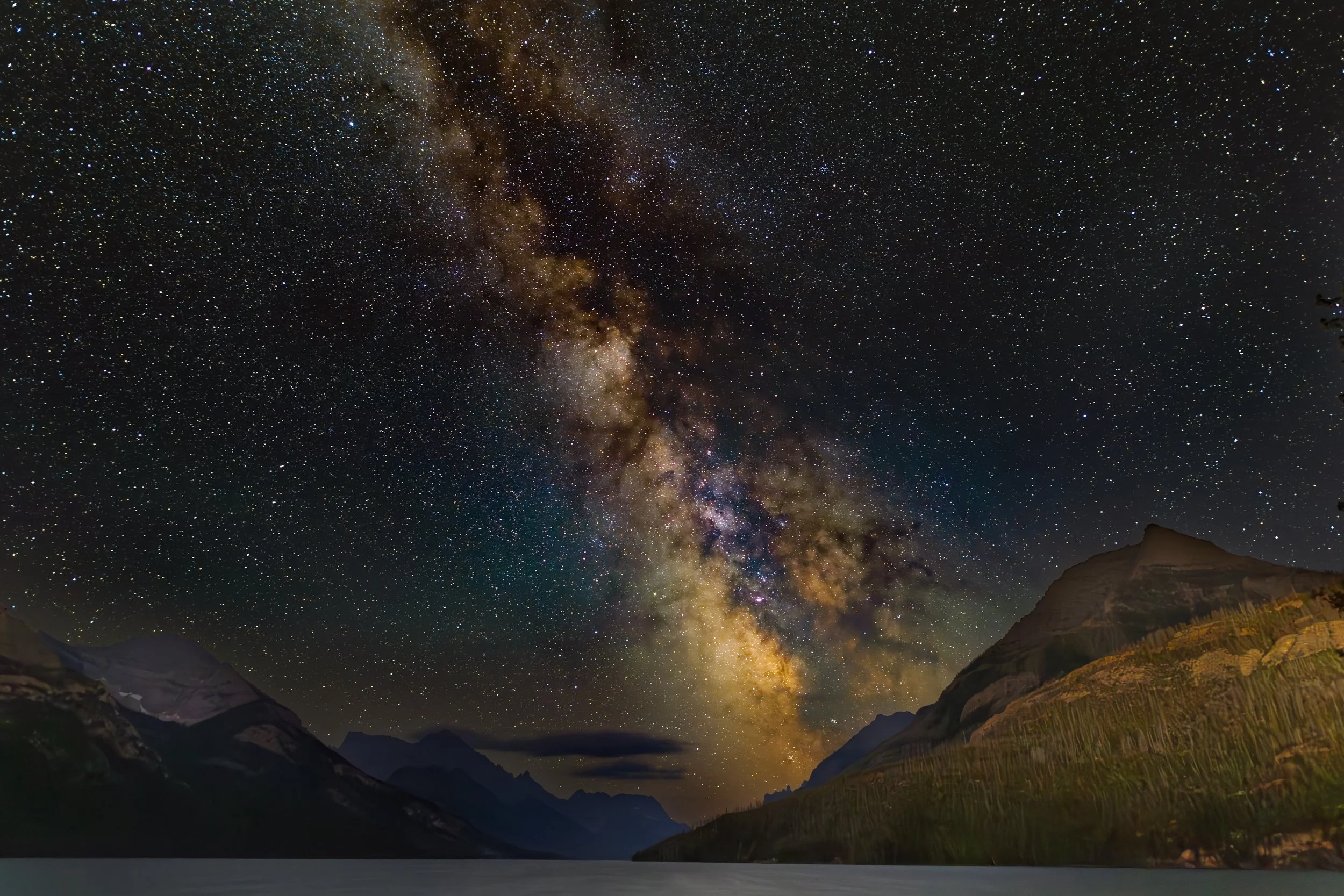 A clear night sky filled with stars and the Milky Way galaxy, with mountains and a body of water in the foreground.