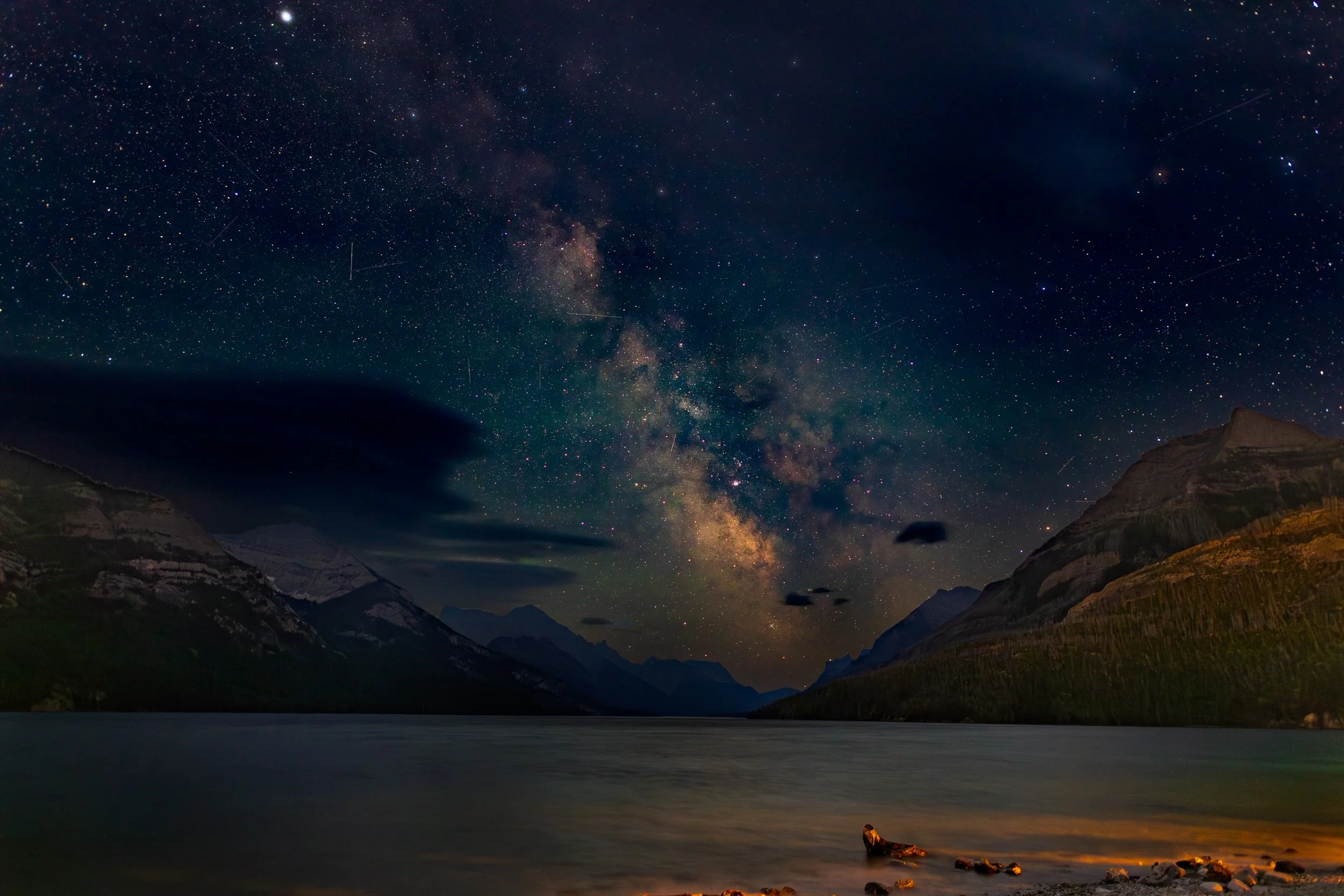 Night sky filled with stars and the Milky Way galaxy above a lake with mountains on either side. A few clouds and rocks are visible along the shoreline.