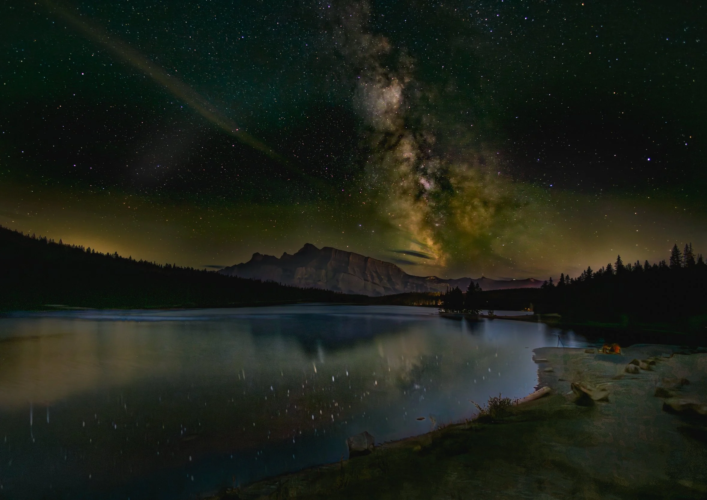 Nighttime landscape with a star-filled sky, showcasing the Milky Way galaxy, over a lake surrounded by forest and mountains.