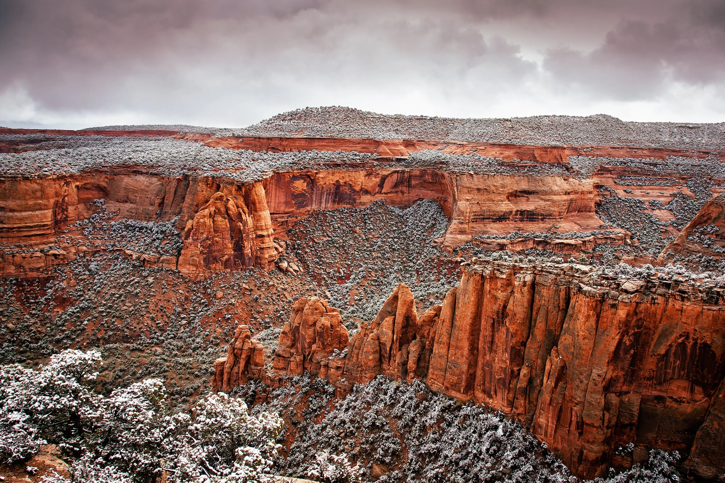 Snow-covered red rock formations and cliffs in a canyon under cloudy sky.