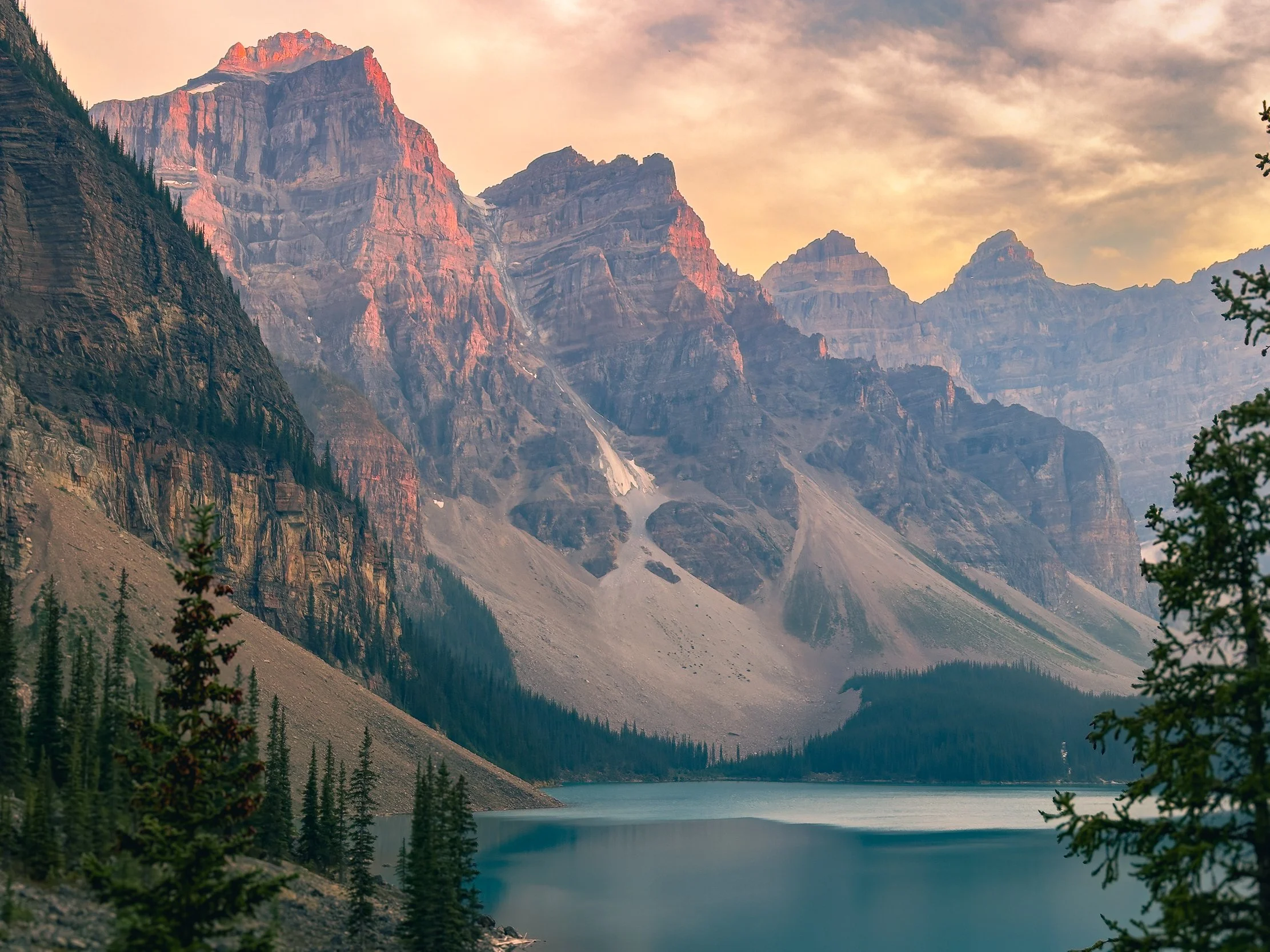 A scenic view of a mountain range with peaks bathed in a warm light, towering over a forested area and a calm lake below.