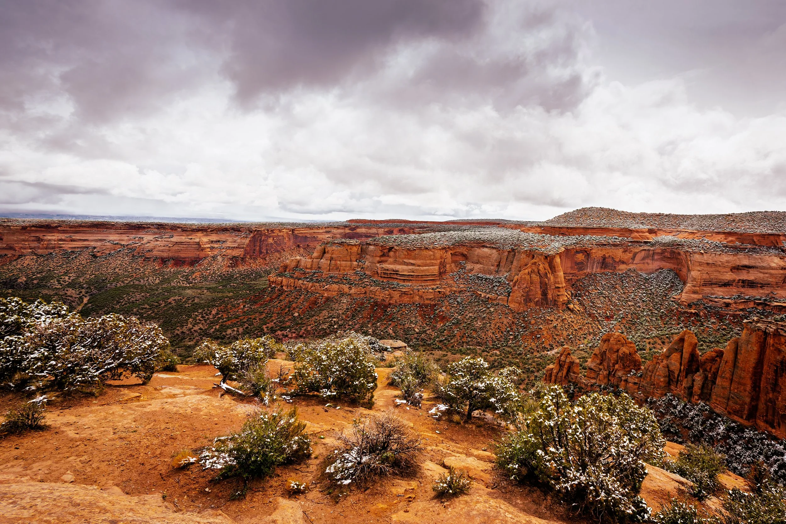 A vast canyon landscape with reddish-brown cliffs and sparse vegetation, dusted with snow under a cloudy sky.