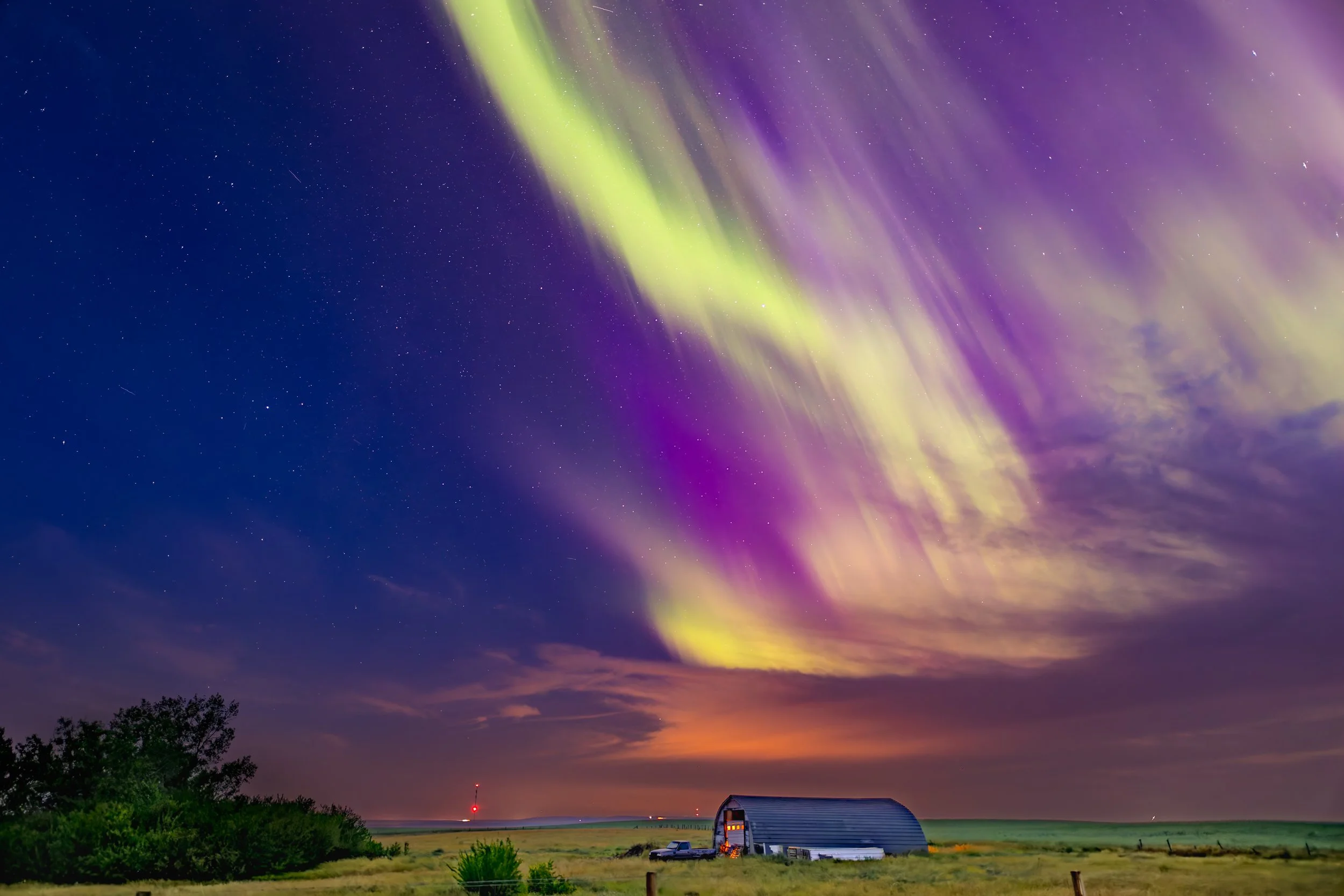 Colorful northern lights display in the night sky over a rural landscape with a barn, a tree, and a car.