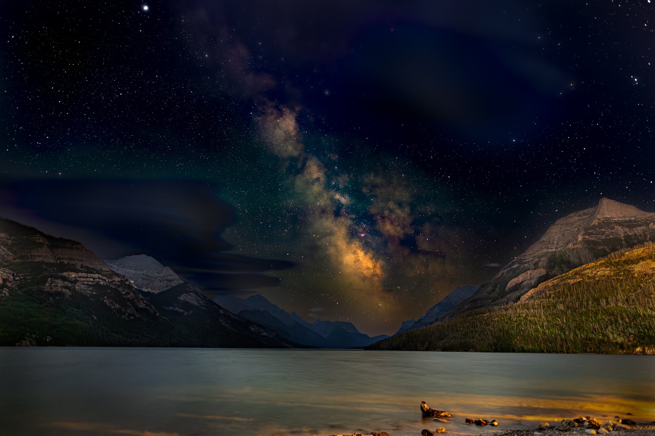 Night sky with the Milky Way galaxy over mountains and a calm lake, with rocks in the foreground.