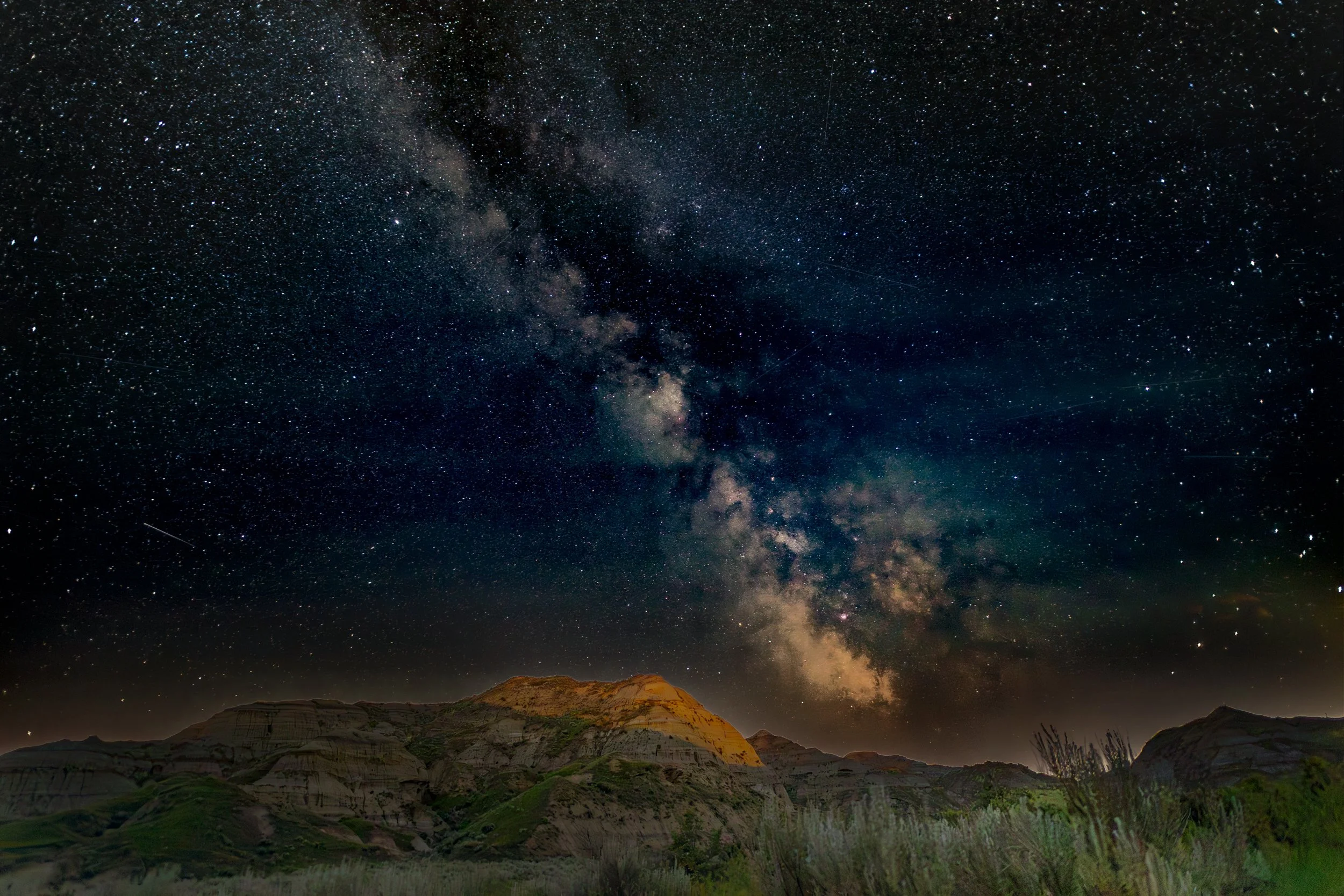 Night sky with the Milky Way galaxy visible over a landscape of mountains and grassy foreground.