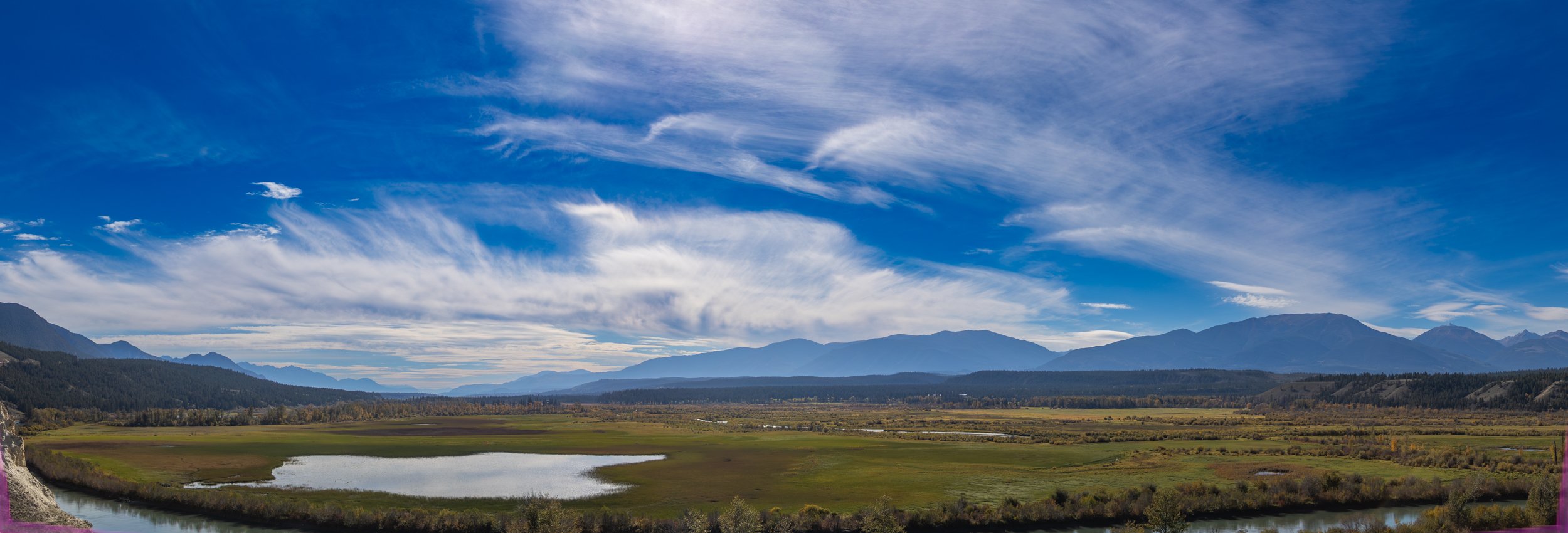 Scenic landscape with a blue sky, wispy clouds, mountains in the distance, a valley with green fields, and a small body of water in the foreground.