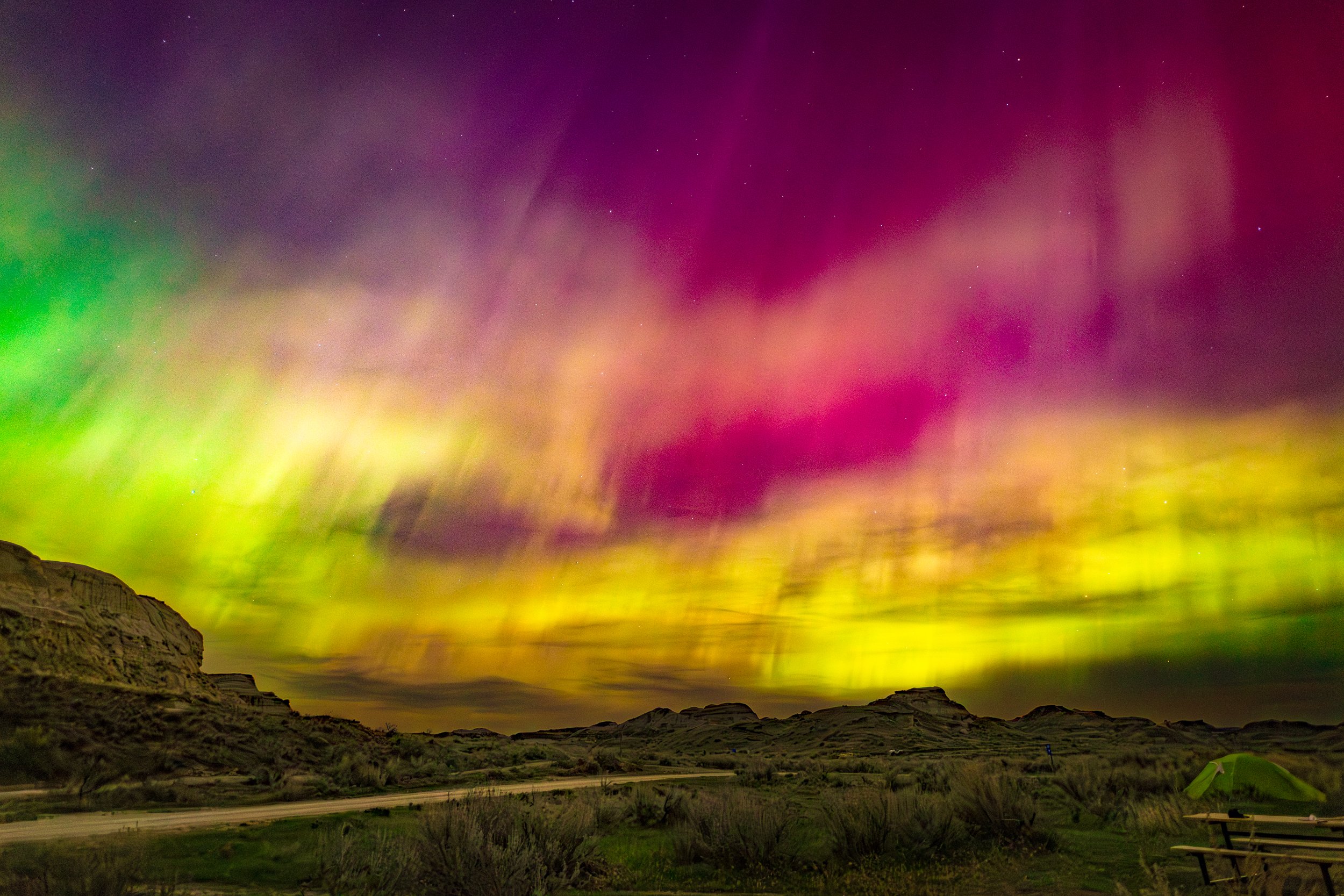Colorful northern lights over a desert landscape with hills, bushes, a tent, and a picnic table.