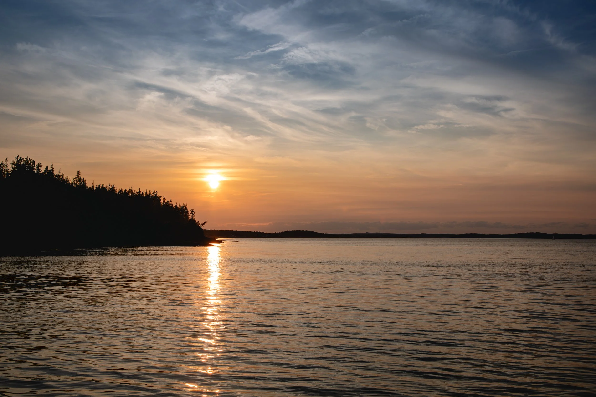 Sunset over a calm body of water with a silhouette of a treeline on the left and a partly cloudy sky.