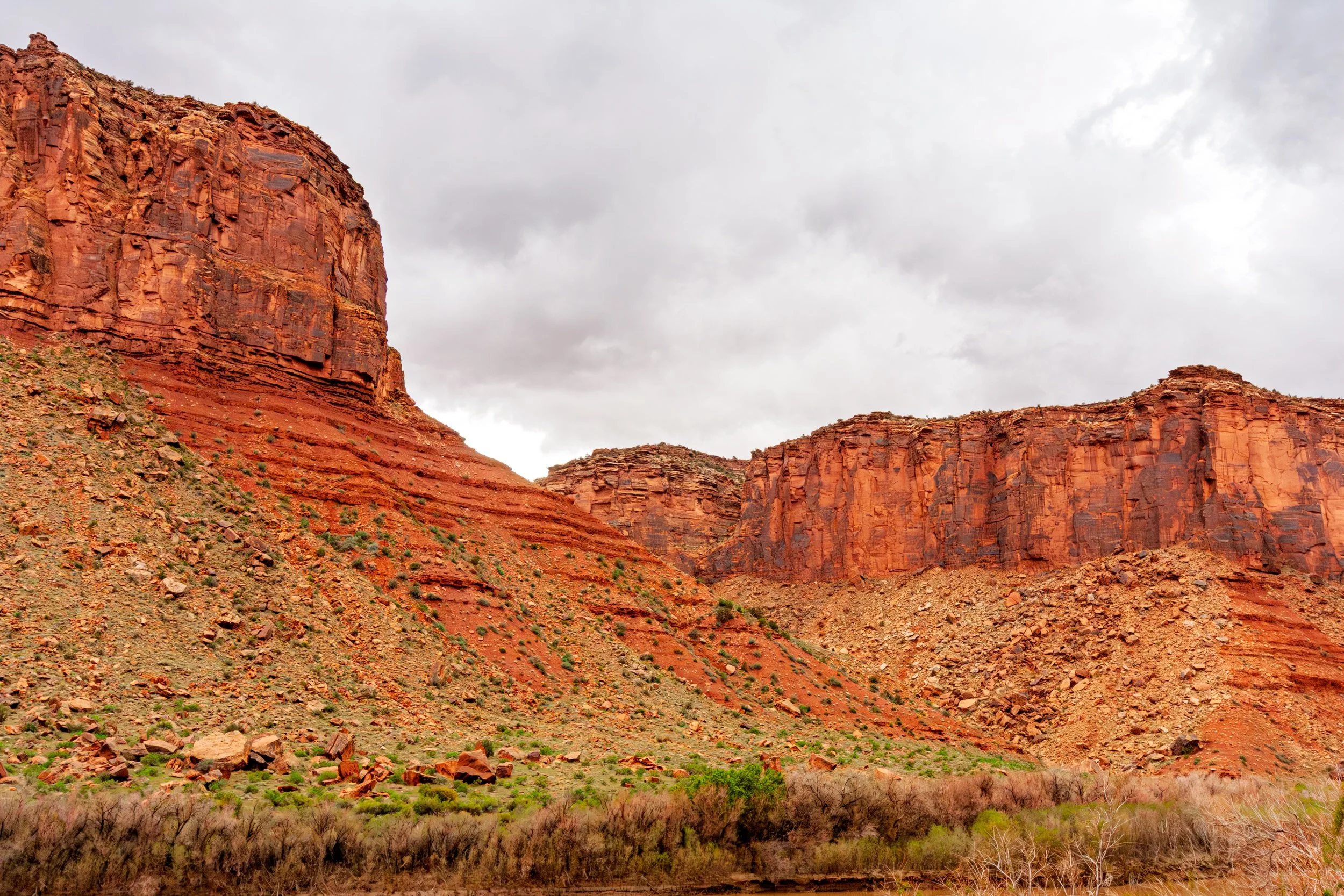 Red rock formations and cliffs in a desert landscape with sparse vegetation and cloudy sky.
