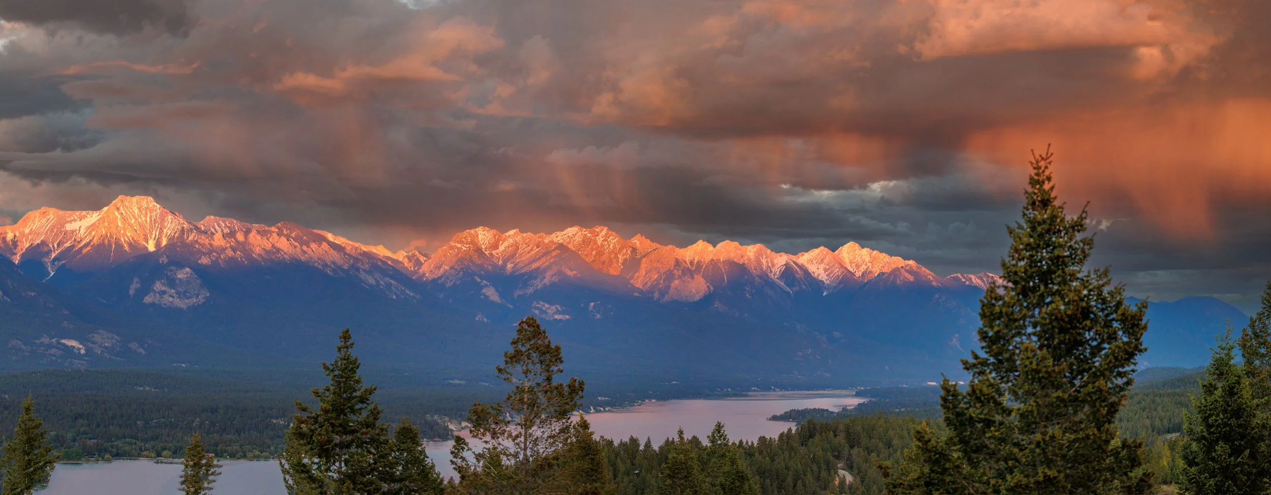 Sunset over teh Columbia Valley with snow-capped mountains with dark clouds and tall trees in the foreground.