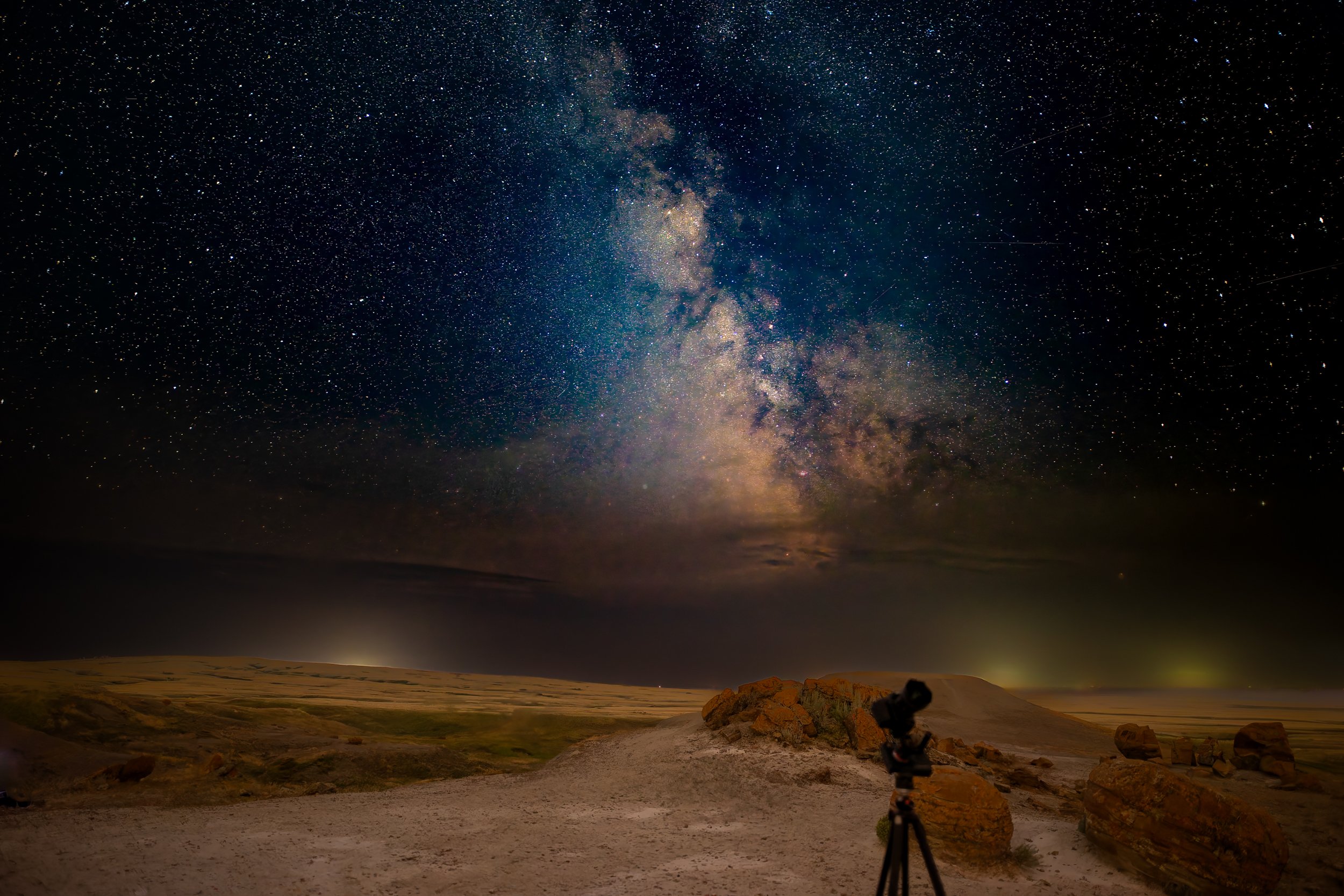 A night sky filled with stars and the Milky Way galaxy, over a desert landscape with rocks and a camera on a tripod in the foreground.