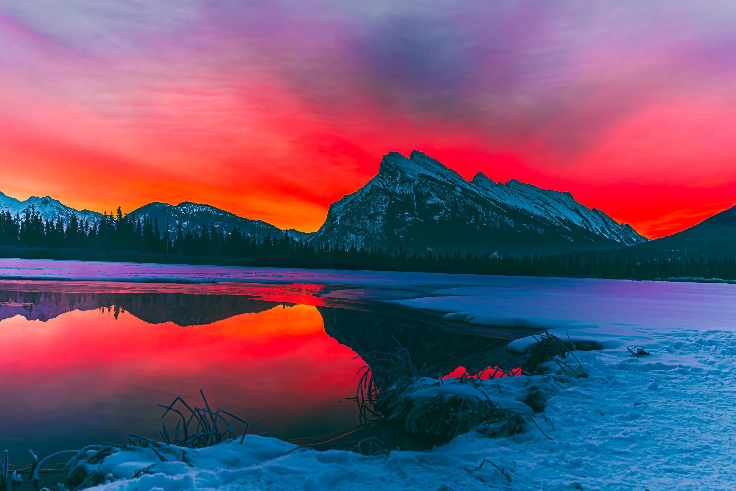 Snow-covered landscape with a partially frozen lake, a mountain with snow, and a colorful sunset sky.