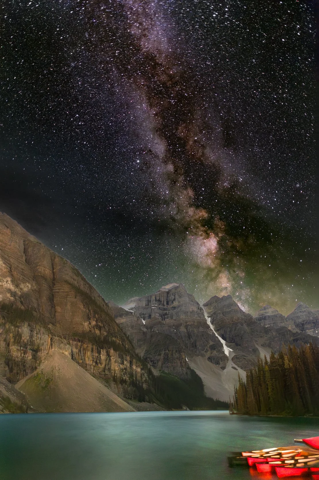 Nighttime view of a starry sky with the Milky Way galaxy over mountains and a body of water, with a small dock and red boats in the foreground.