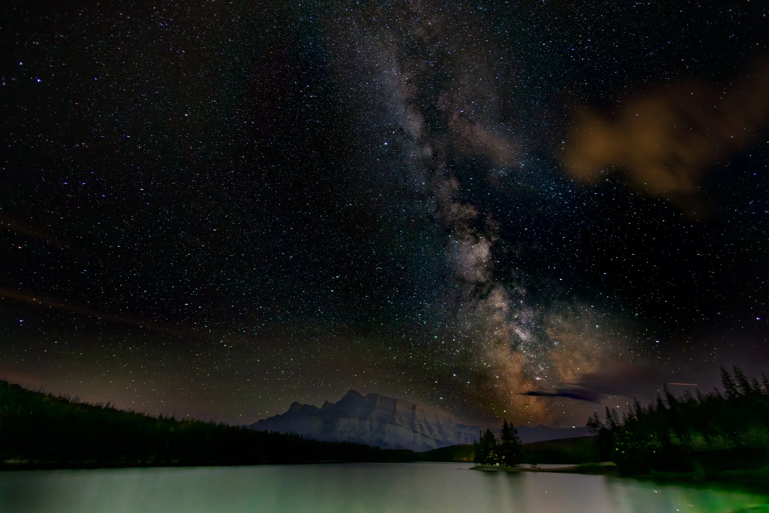 Night sky filled with stars and the Milky Way galaxy over a lake with mountains and forested shoreline.