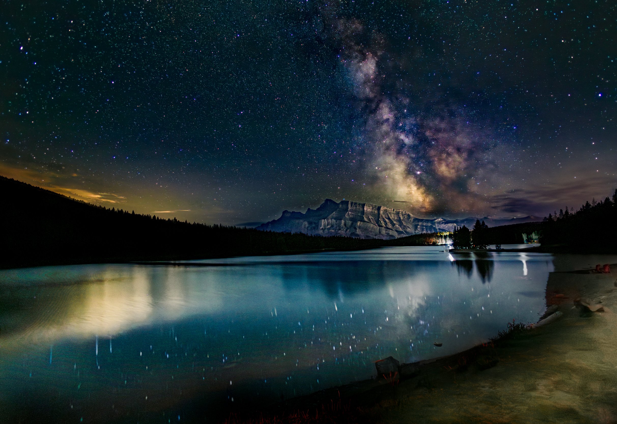 Nighttime scene of a starry sky with the Milky Way galaxy over a mountain range, reflecting in a calm lake with a forested shoreline.