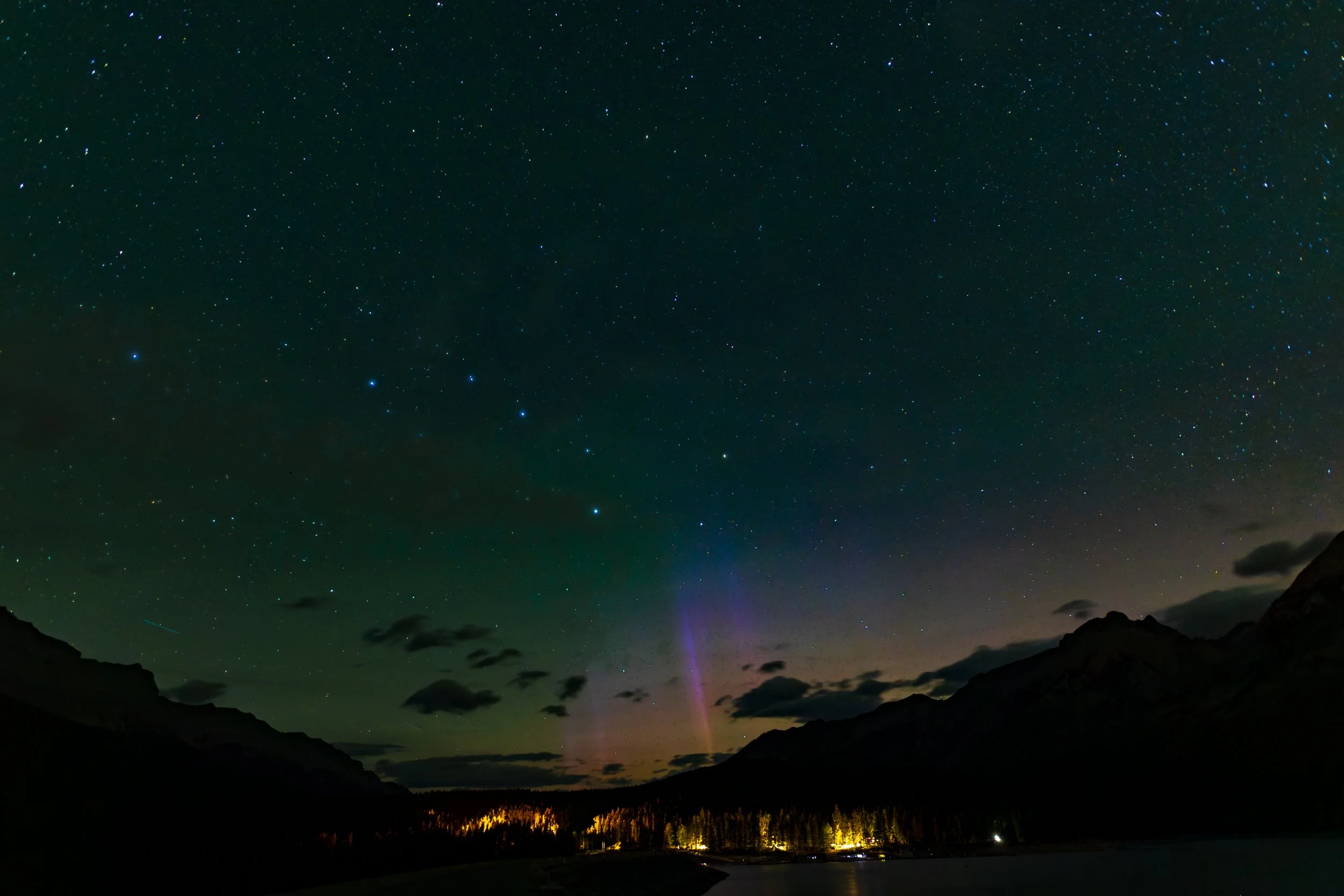 Night sky filled with stars, with mountain silhouettes and a forested area illuminated by lights at the horizon.