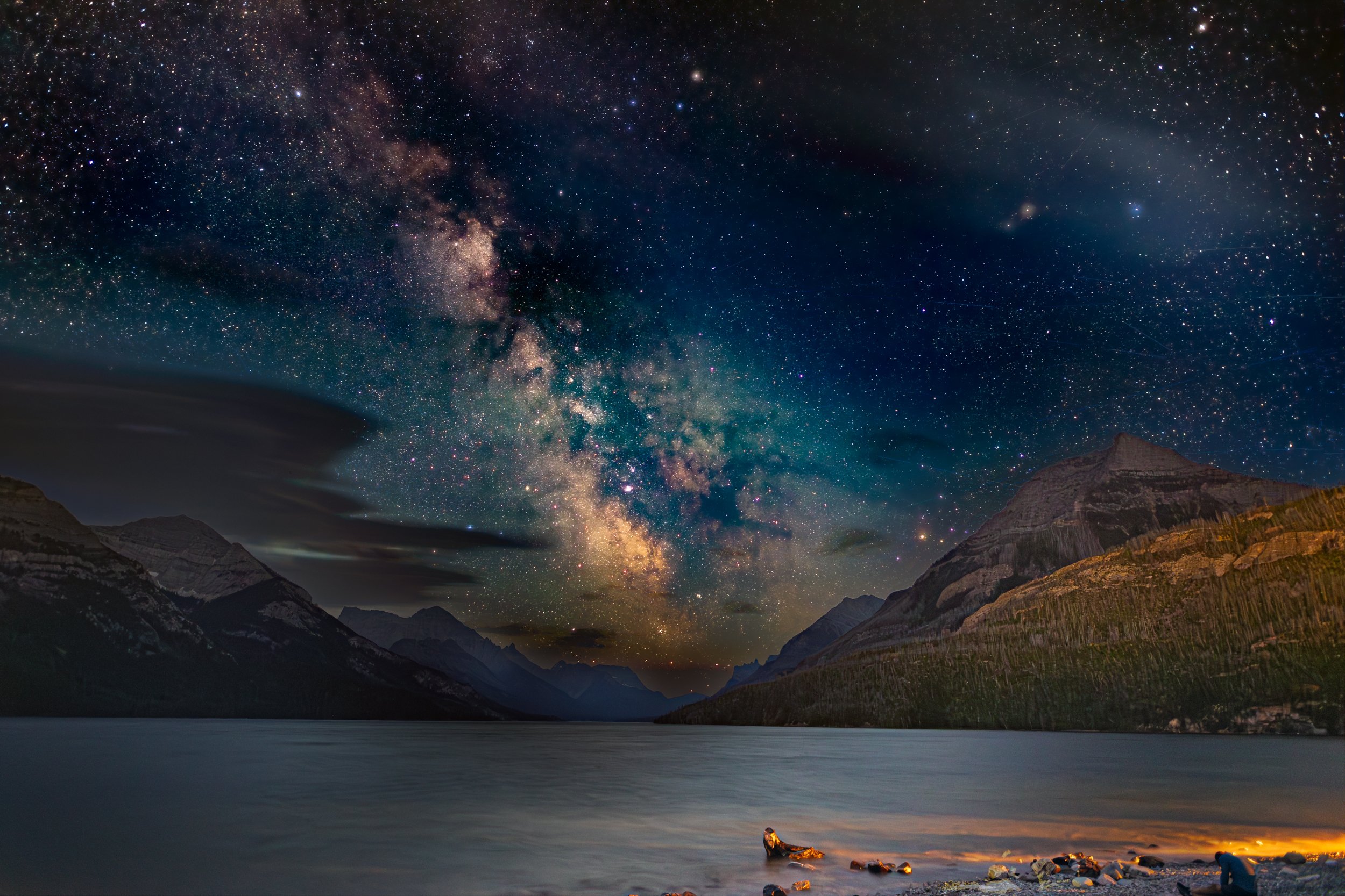 Night sky filled with stars and the Milky Way galaxy above a mountain range and a lake, with a person sitting on the shore.