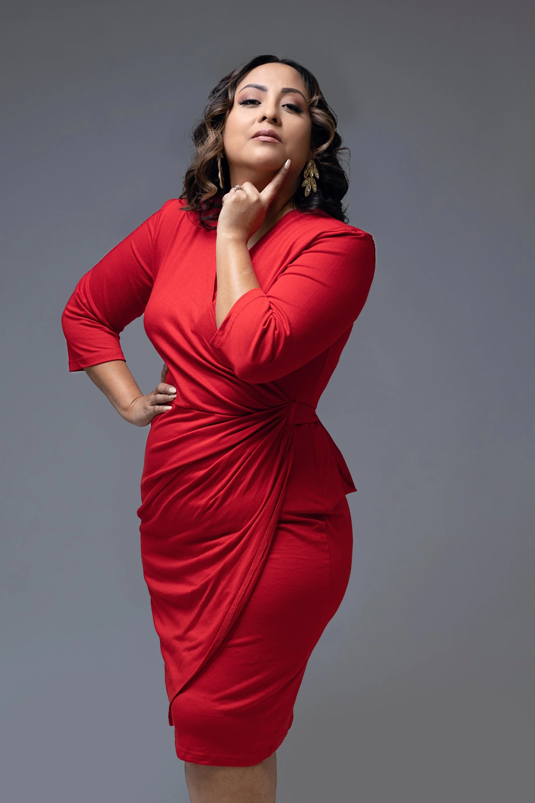 Dark-haired woman in a red dress on a gray background posing for She Means Business Magazine.
