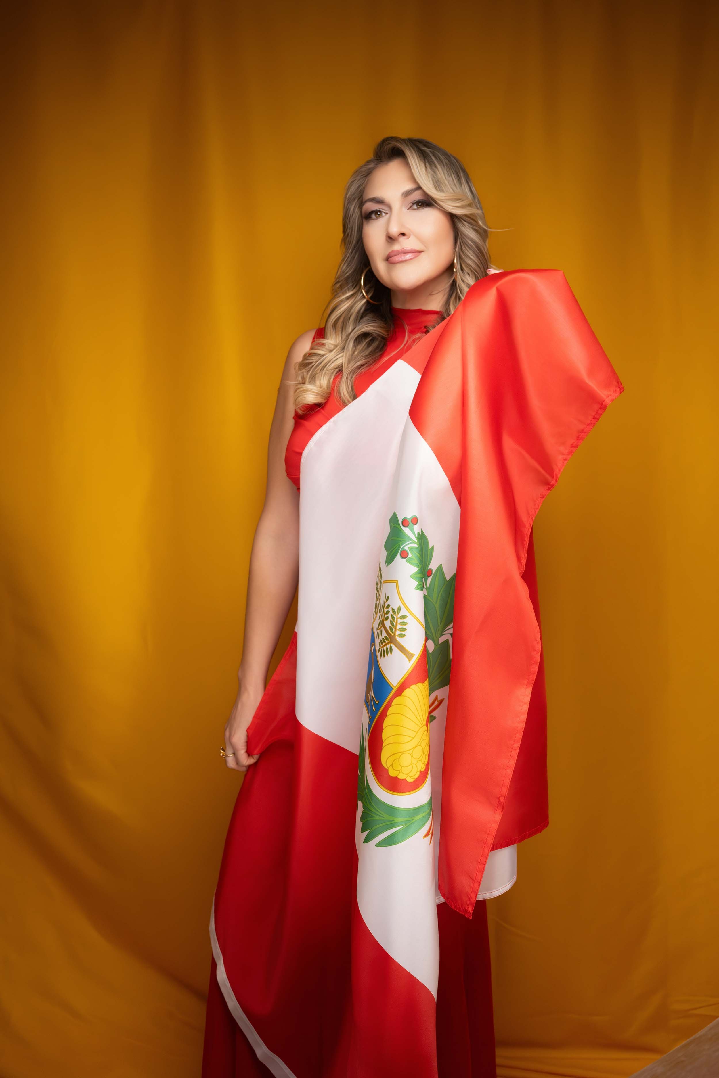 A woman with long blonde wavy hair, wearing gold hoop earrings, dressed in a red dress, draped in a Mexican flag, standing in front of a golden-yellow curtain.