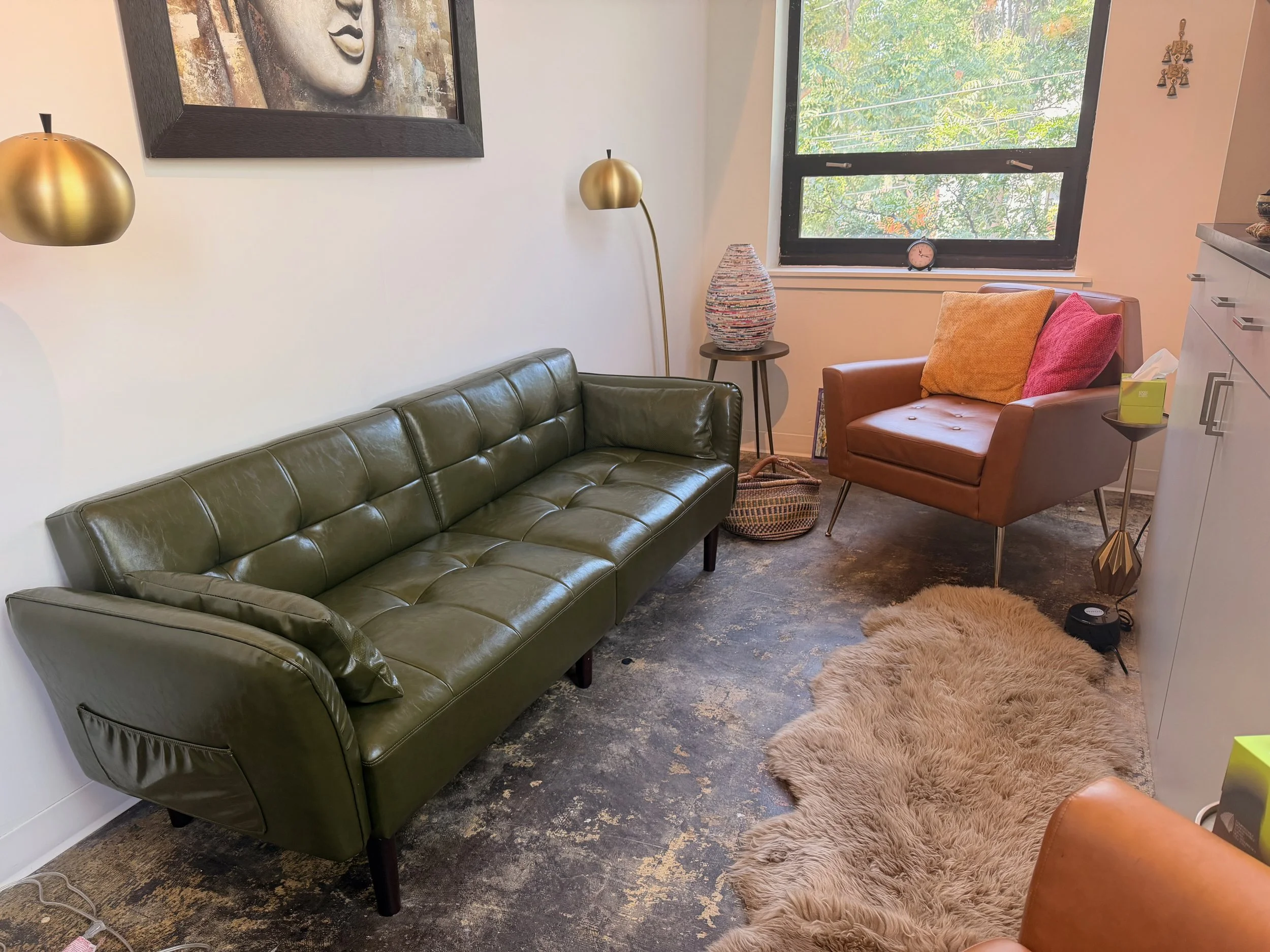 A living room with a green leather sofa, a tan armchair with orange and pink pillows, a shaggy beige rug, a large window with trees outside, a colorful vase on a small table, a floor lamp, and wall decor. There is also a framed artwork of a face on the wall.